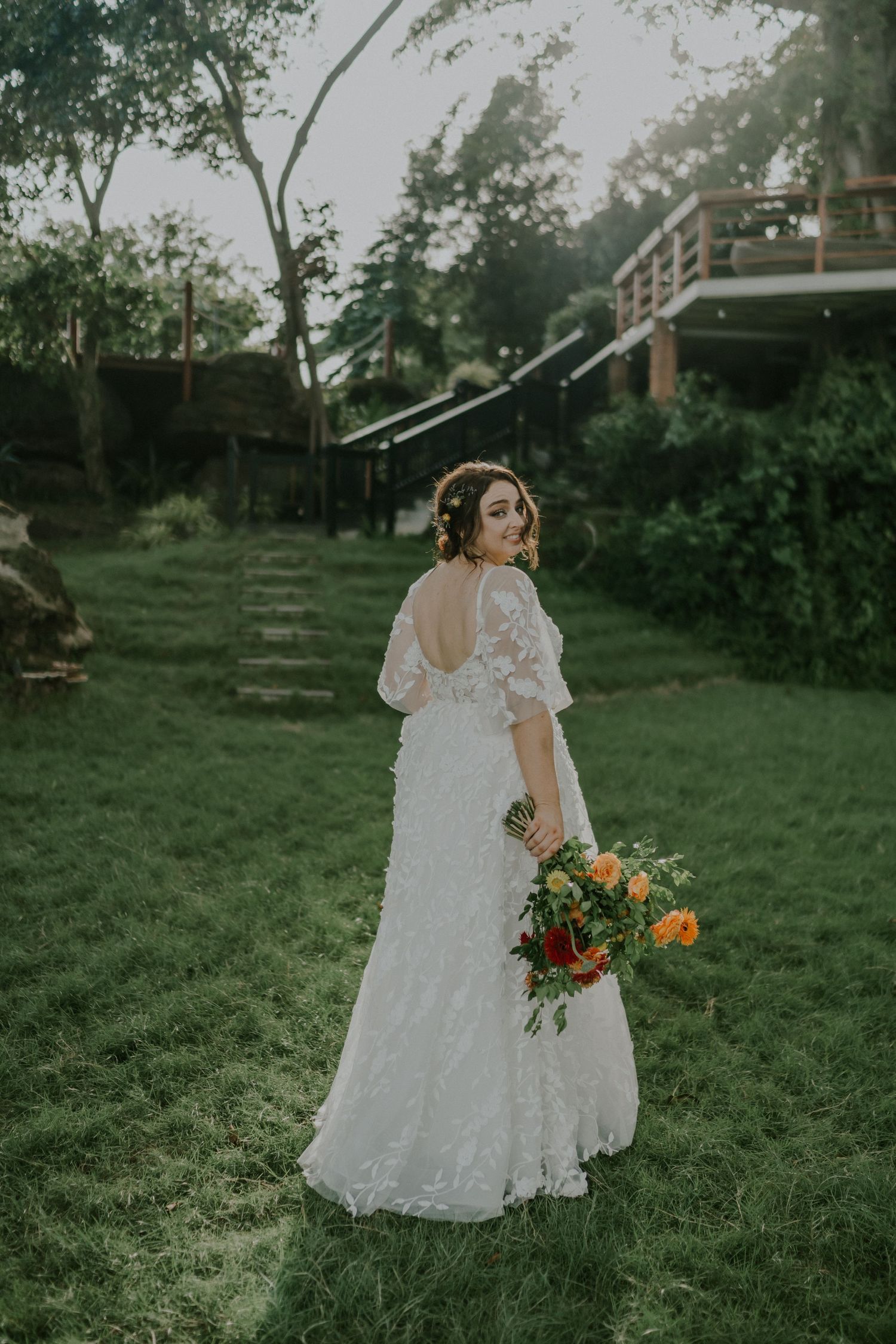 Bride in lace gown poses with colorful bouquet in garden setting with rustic architecture.