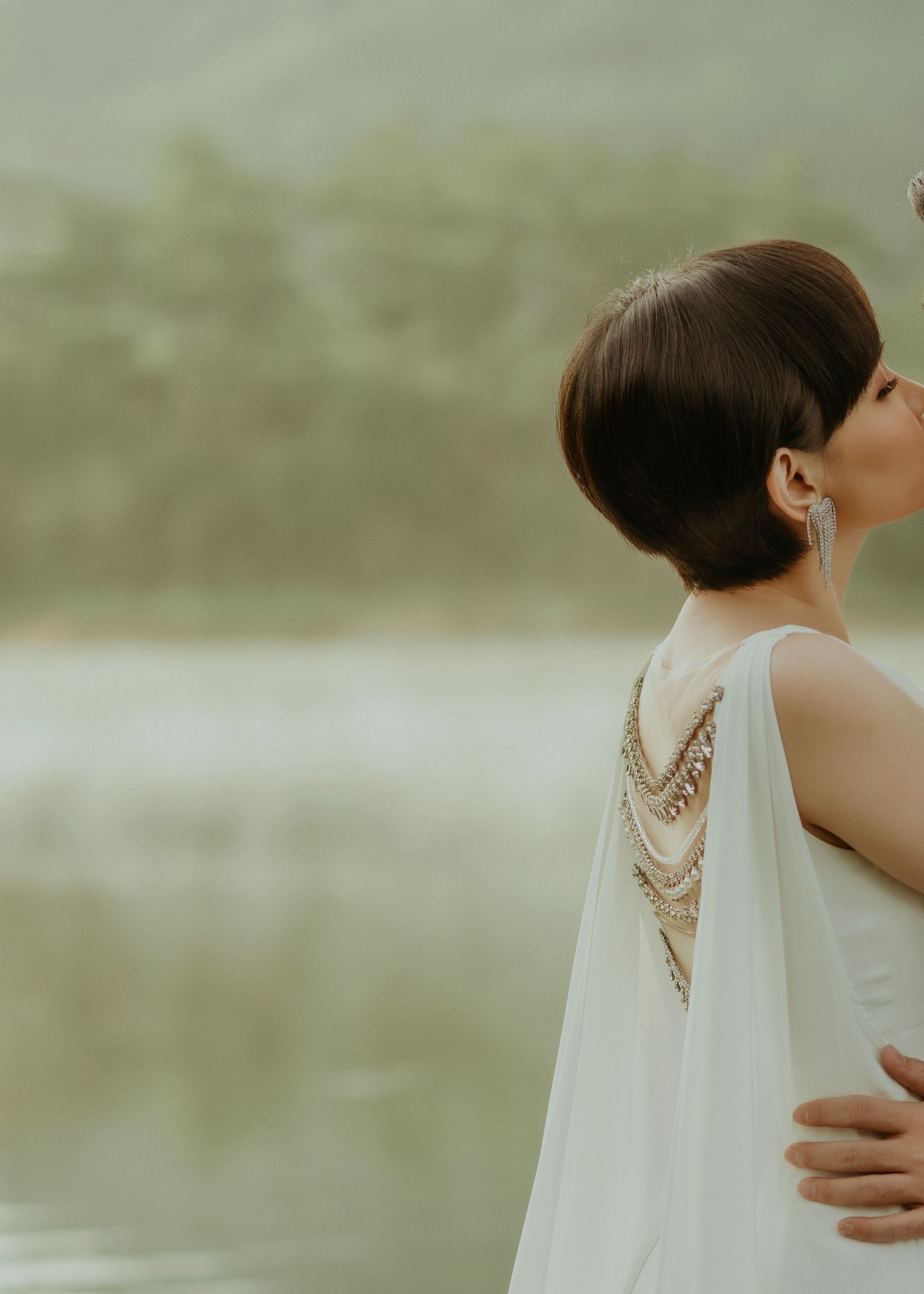 Side profile view of woman in white dress with statement back necklace against blurred natural background.