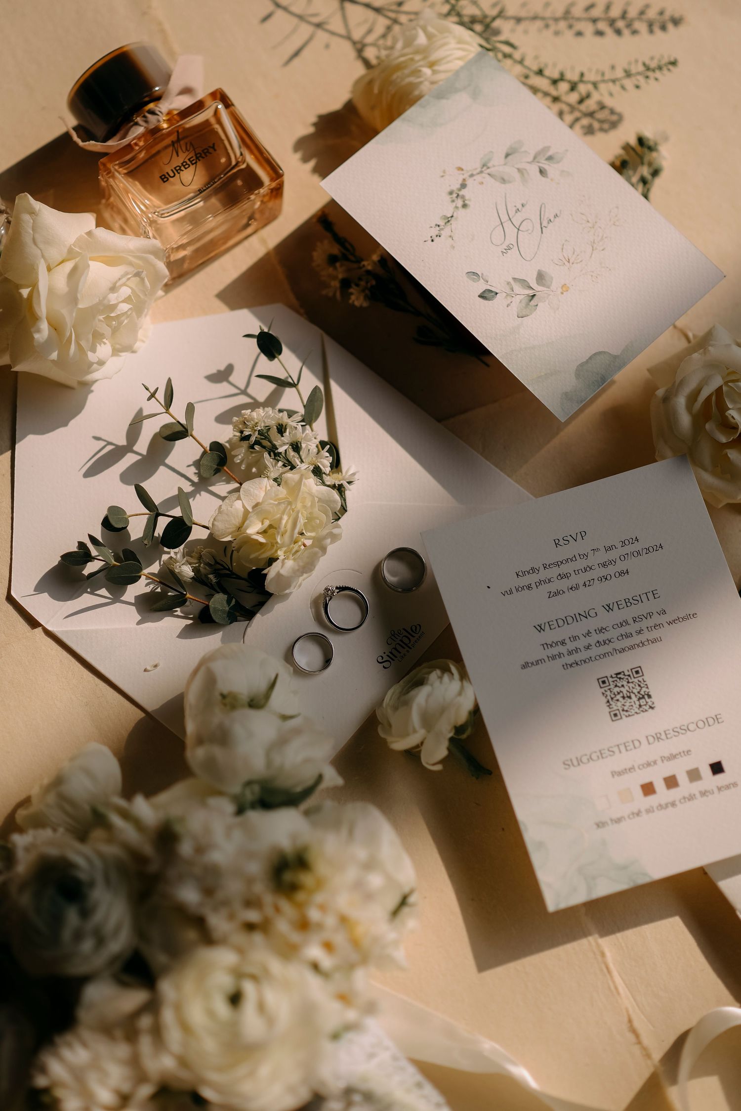 Wedding stationery flatlay with floral details, rings, and perfume in warm sunlight.