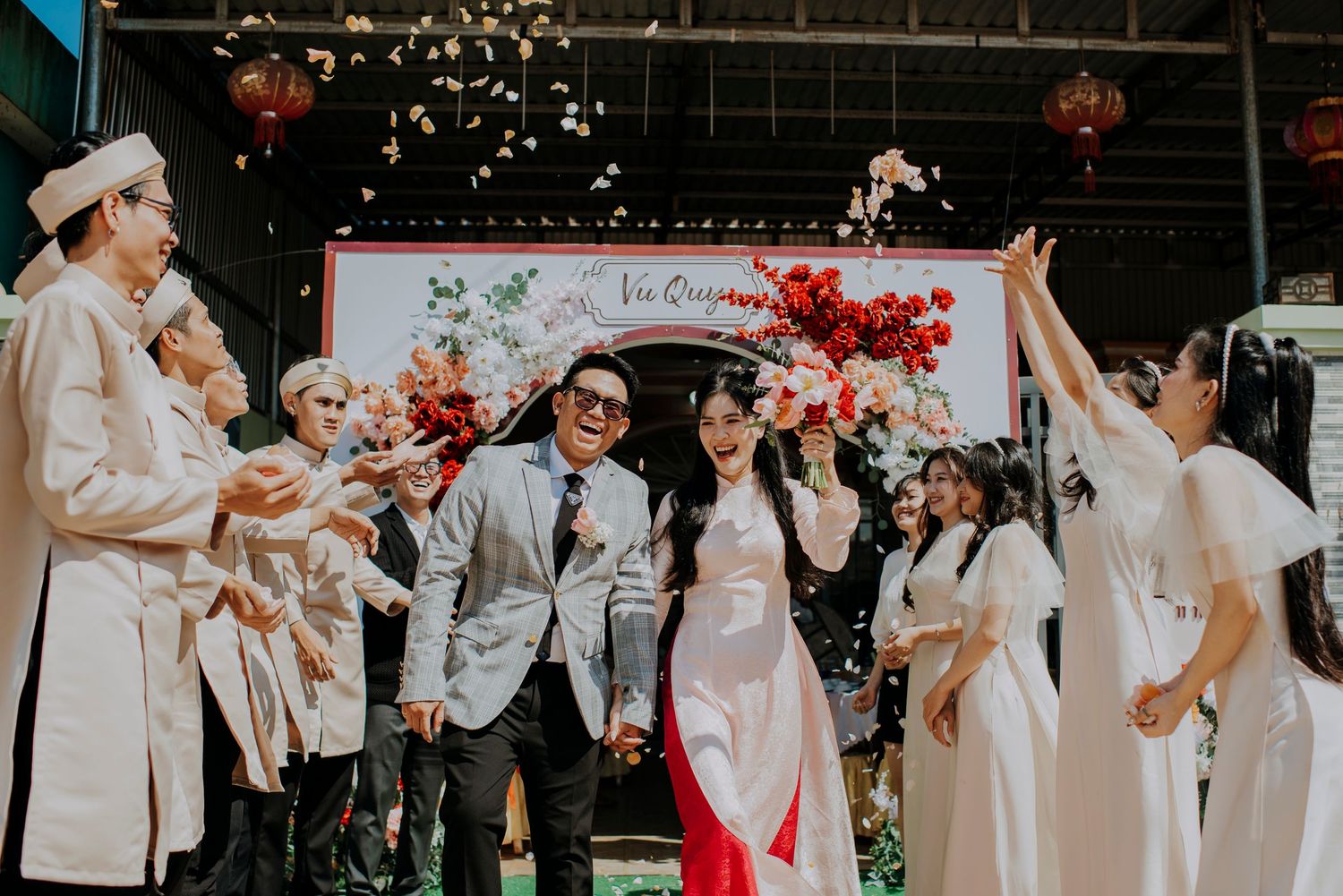 Wedding celebration with guests tossing confetti under red floral arch decoration.