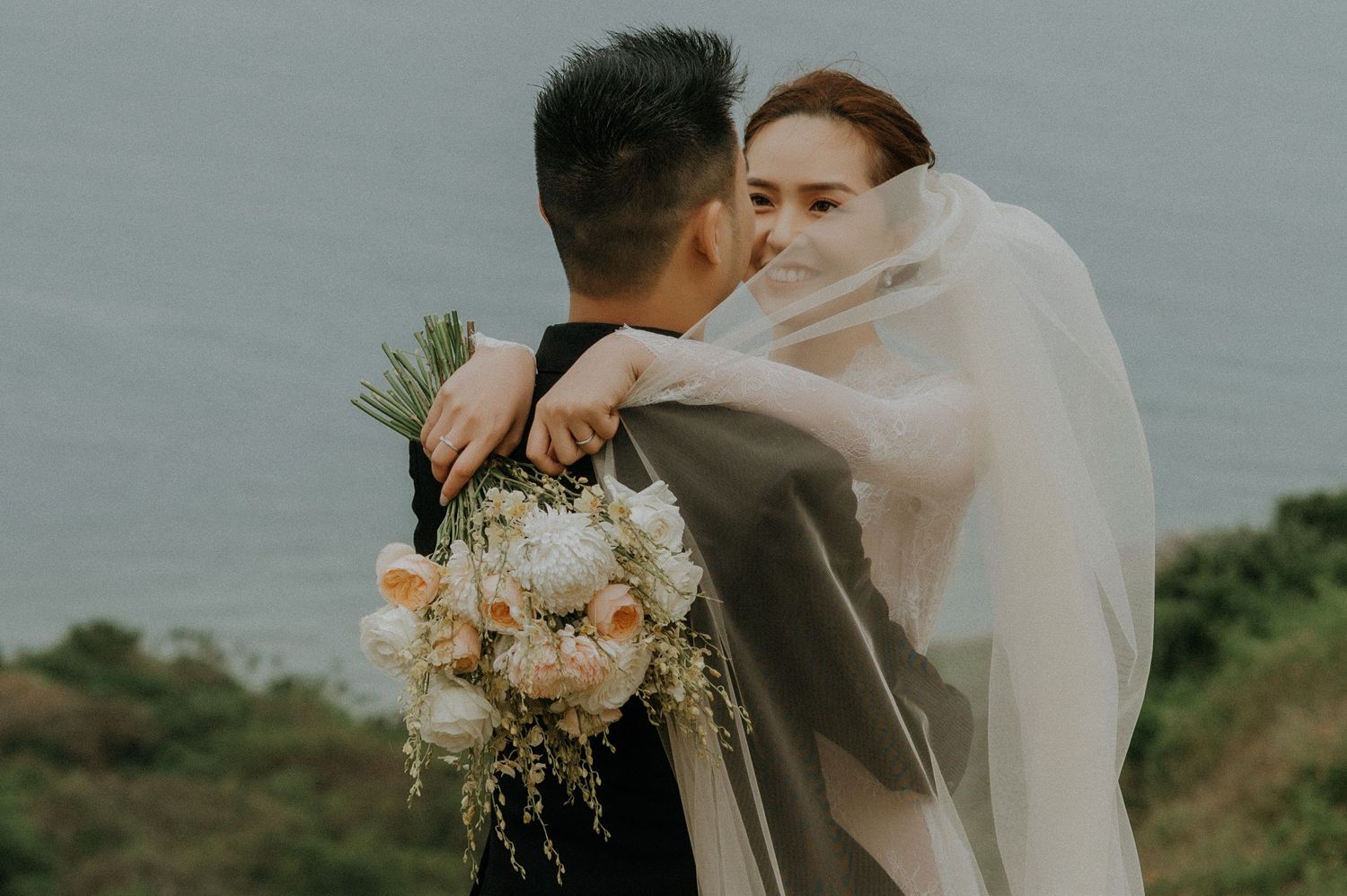 A romantic wedding couple embraces on a cliff overlooking the ocean with a floral bouquet and flowing veil.