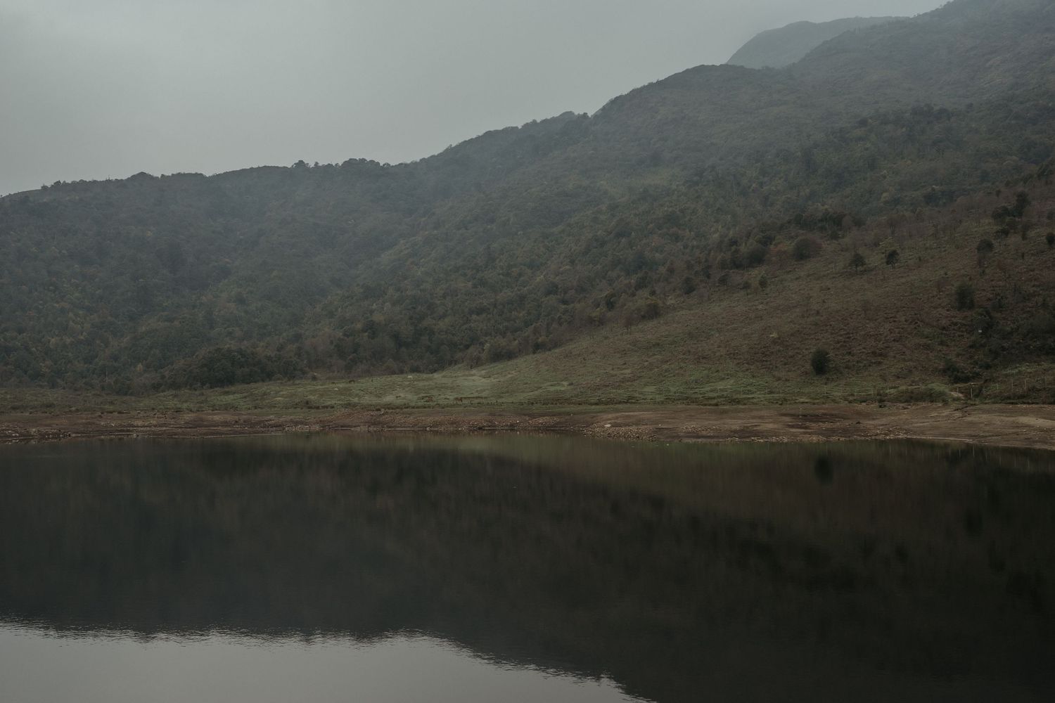 A still lake reflects the surrounding mountains under a moody, overcast sky.