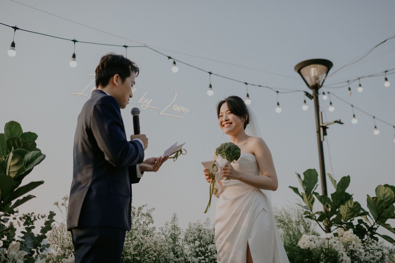 An outdoor evening ceremony under string lights with decorative plants in the background.