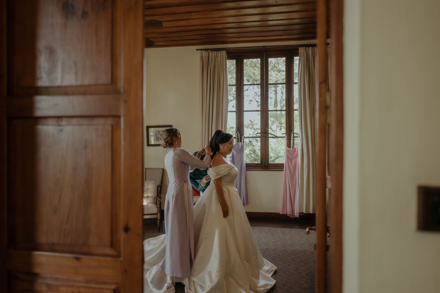 Wedding party helps adjust a flowing white dress in a doorway of rustic room with natural light streaming through window.