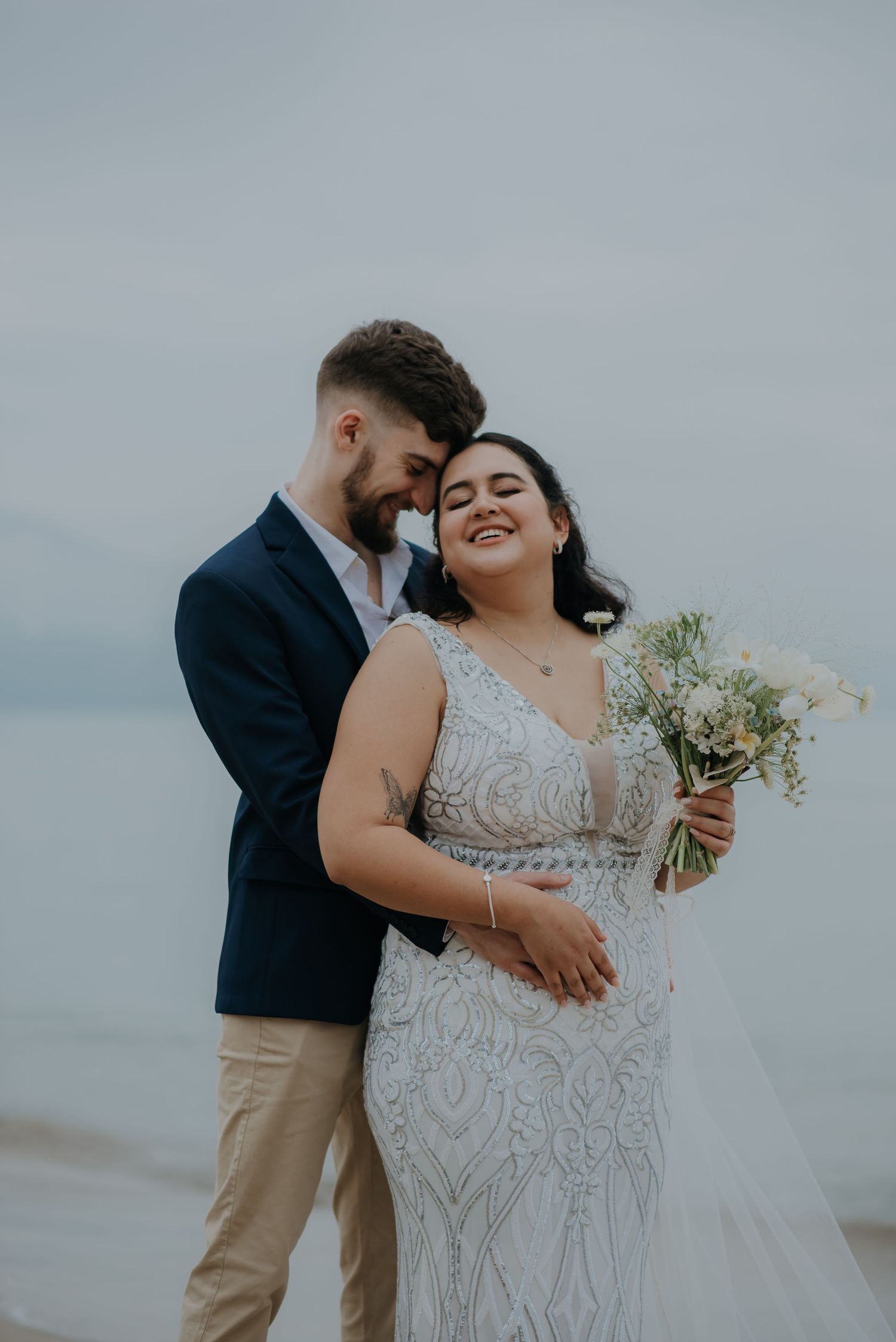 A romantic wedding embrace on a cloudy beach with a white bouquet.