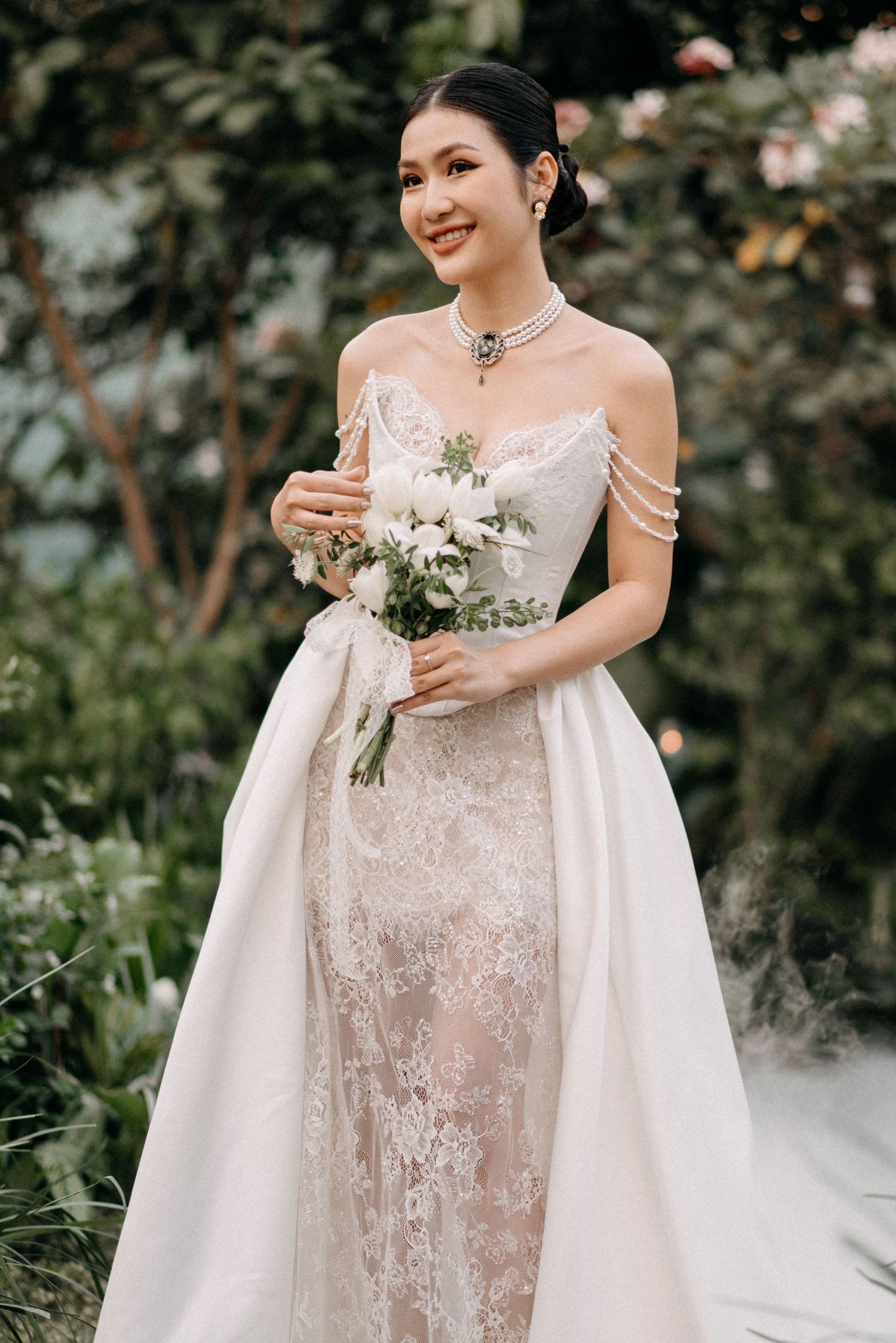 Person in lace wedding gown holds white flower bouquet in garden setting.