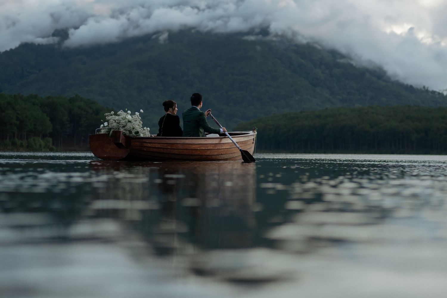Two people sit together in a wooden rowboat on a misty mountain lake surrounded by evergreen forest.