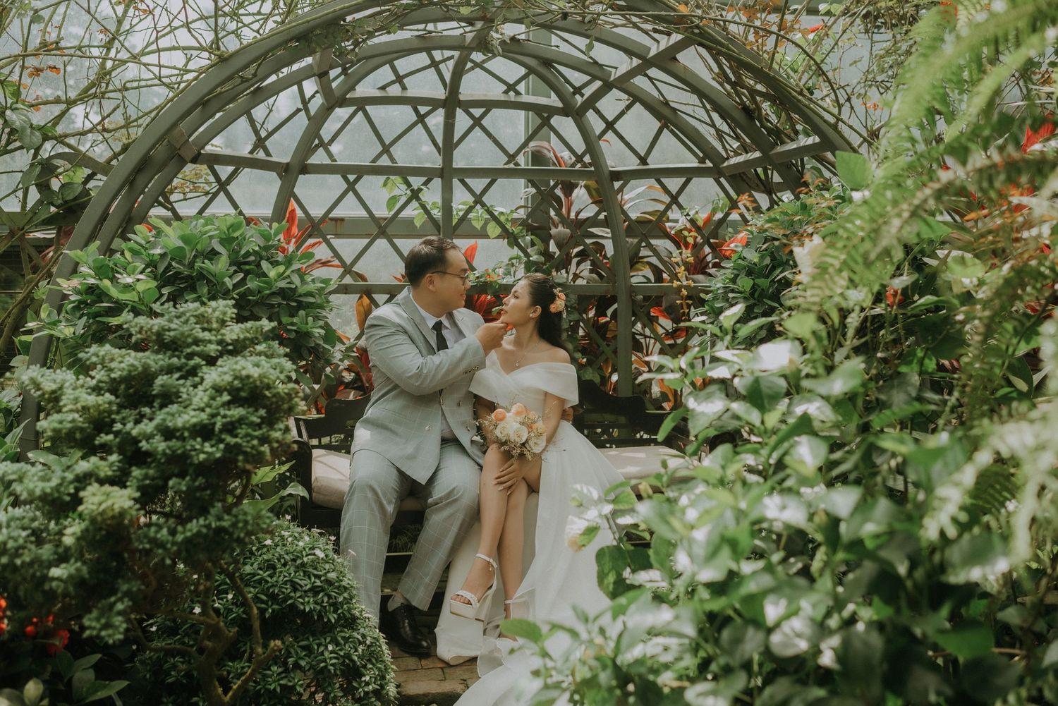 A romantic moment inside a lush greenhouse with arched glass ceiling and blooming plants surrounding a cozy seating area.