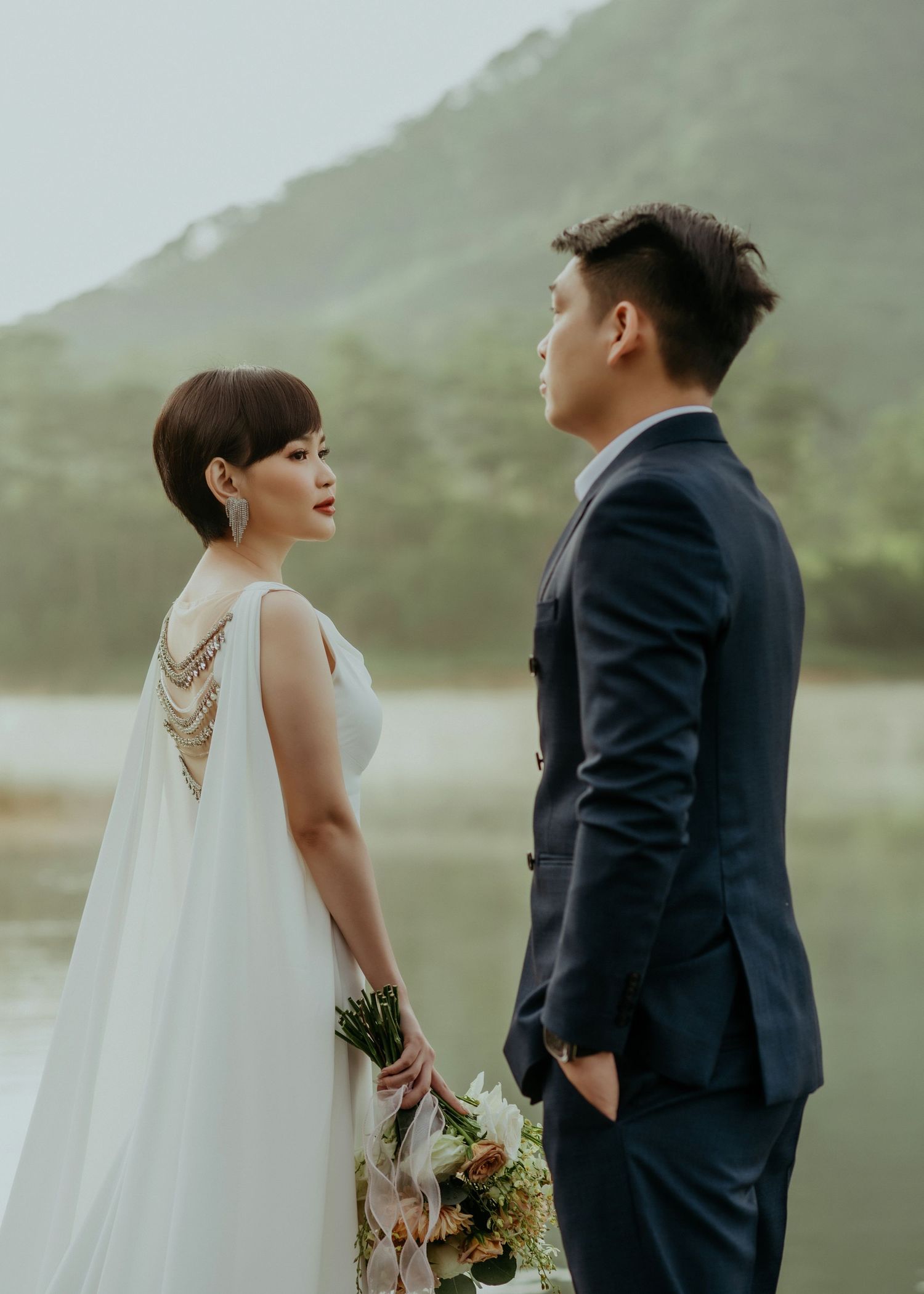 A bride in a flowing white dress with beaded back detail and groom in navy suit pose against misty mountain scenery.