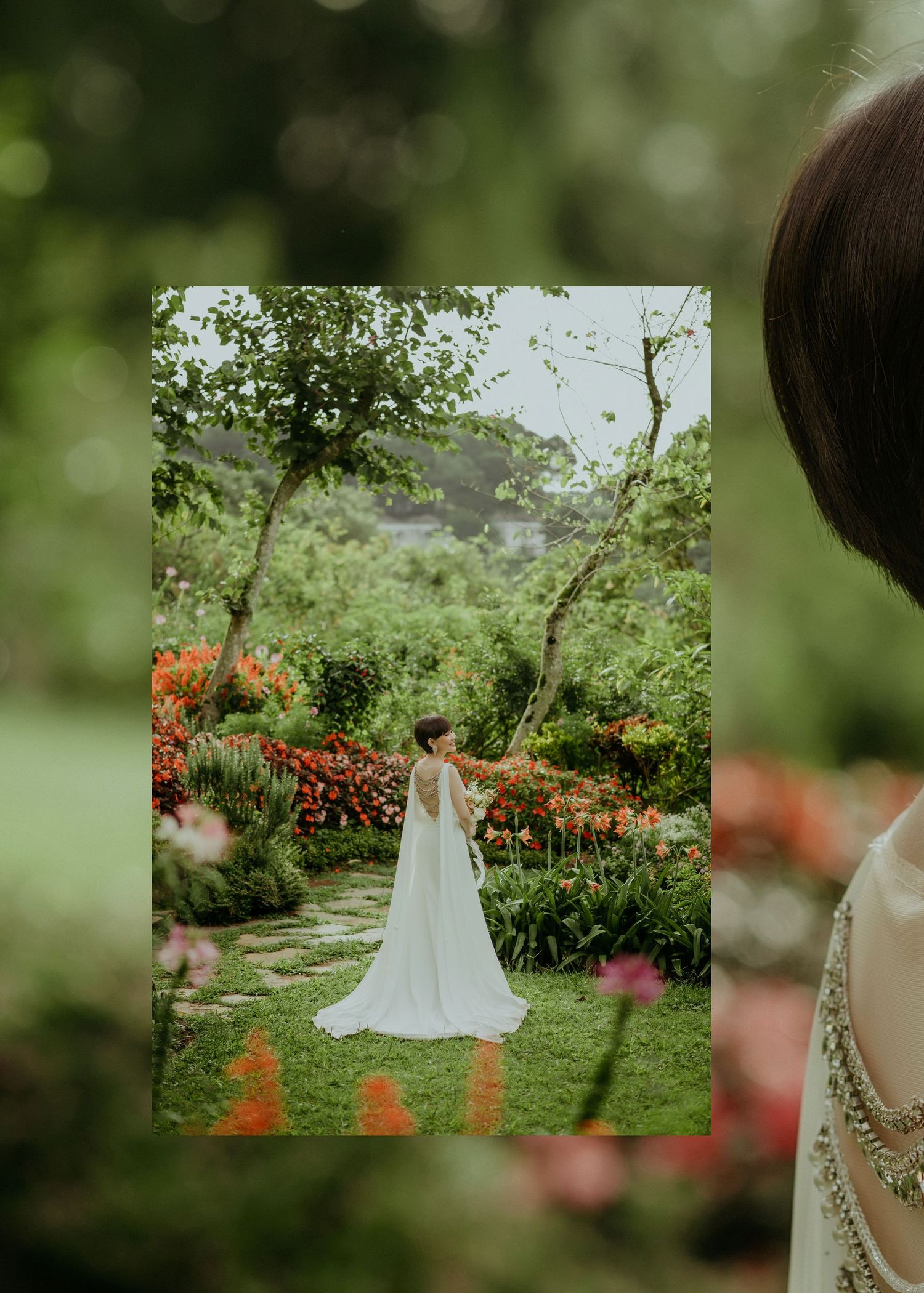 A bride in a flowing white wedding gown stands in a lush garden path surrounded by vibrant orange flowers and greenery.