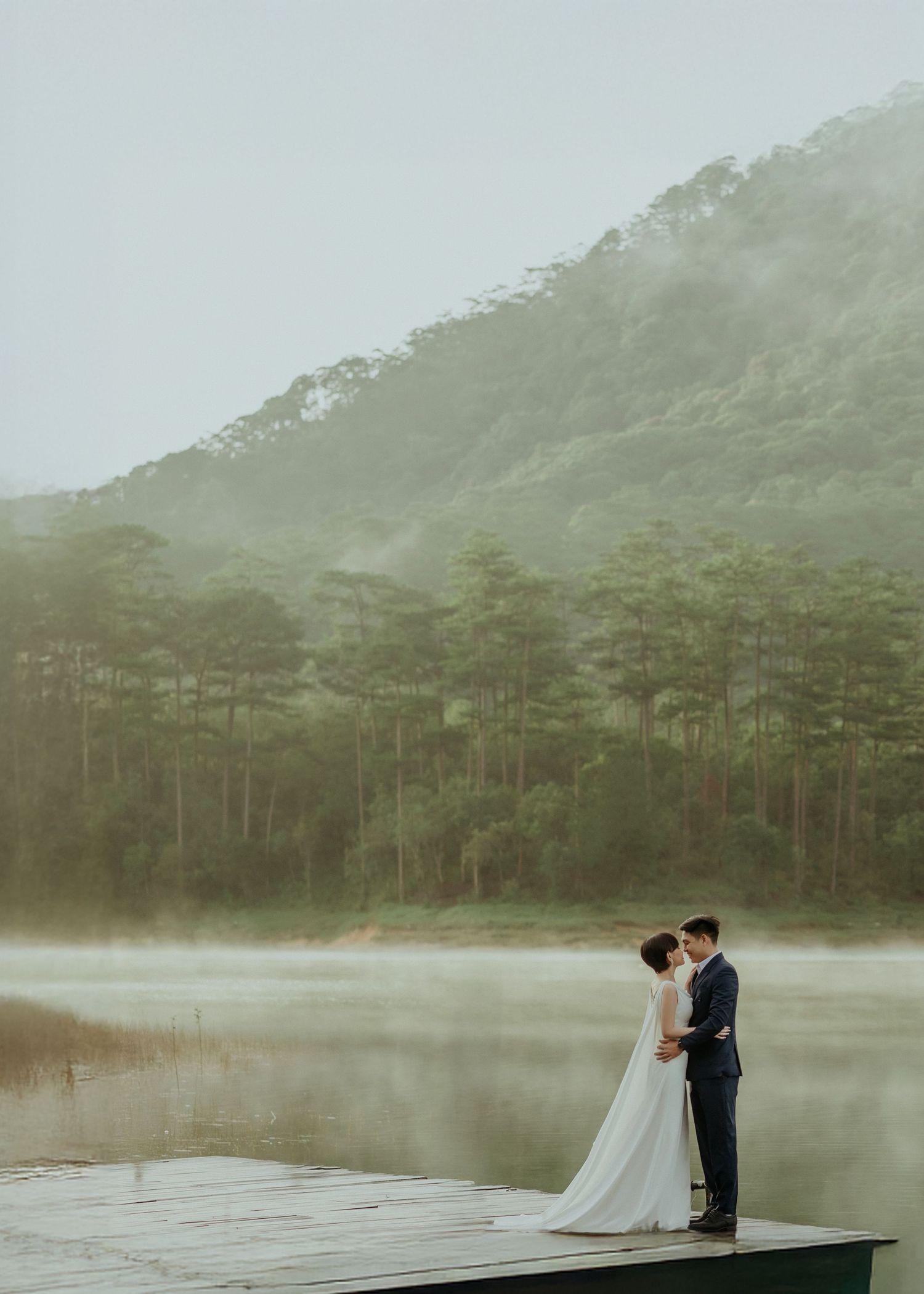 Couple stands together on misty lake dock with foggy mountains and pine trees creating ethereal romantic atmosphere.