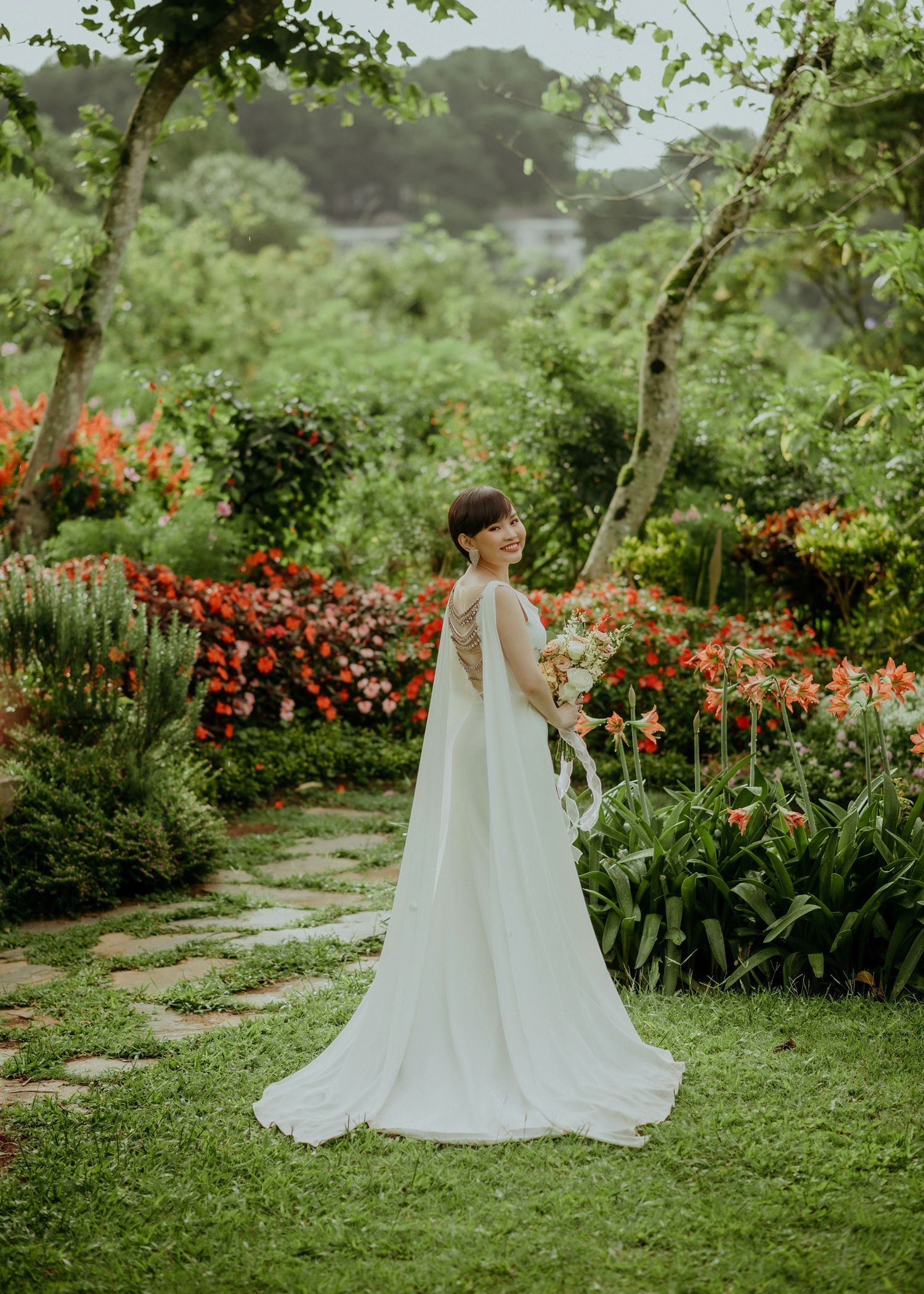 Elegant white wedding gown with flowing cape photographed in a lush garden filled with vibrant orange flowers and greenery.