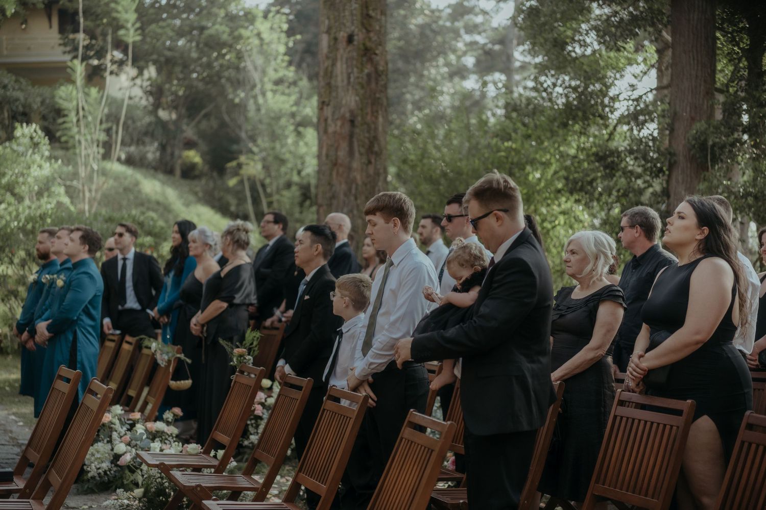 A group of formally dressed musicians perform with wooden djembe drums in an outdoor forest setting.