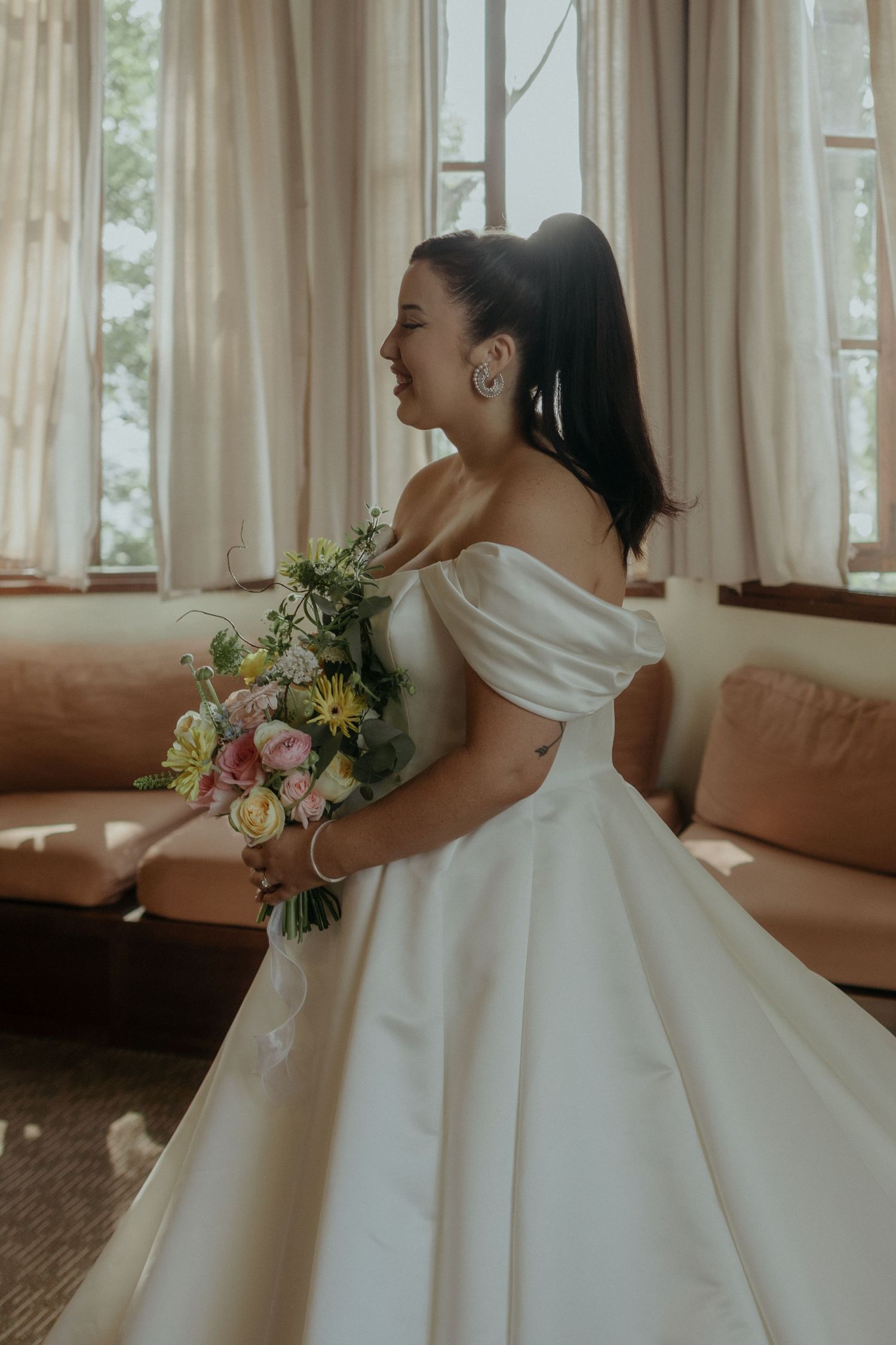 A side view of a bride in a classic white ball gown holding a colorful bouquet in a sunlit room.