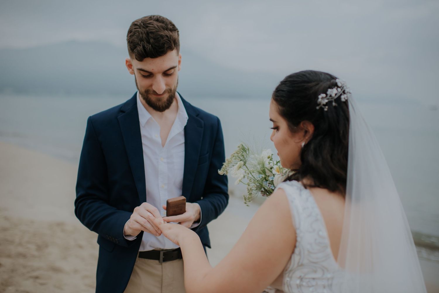 A couple exchanges rings during their beach wedding ceremony under an overcast sky.