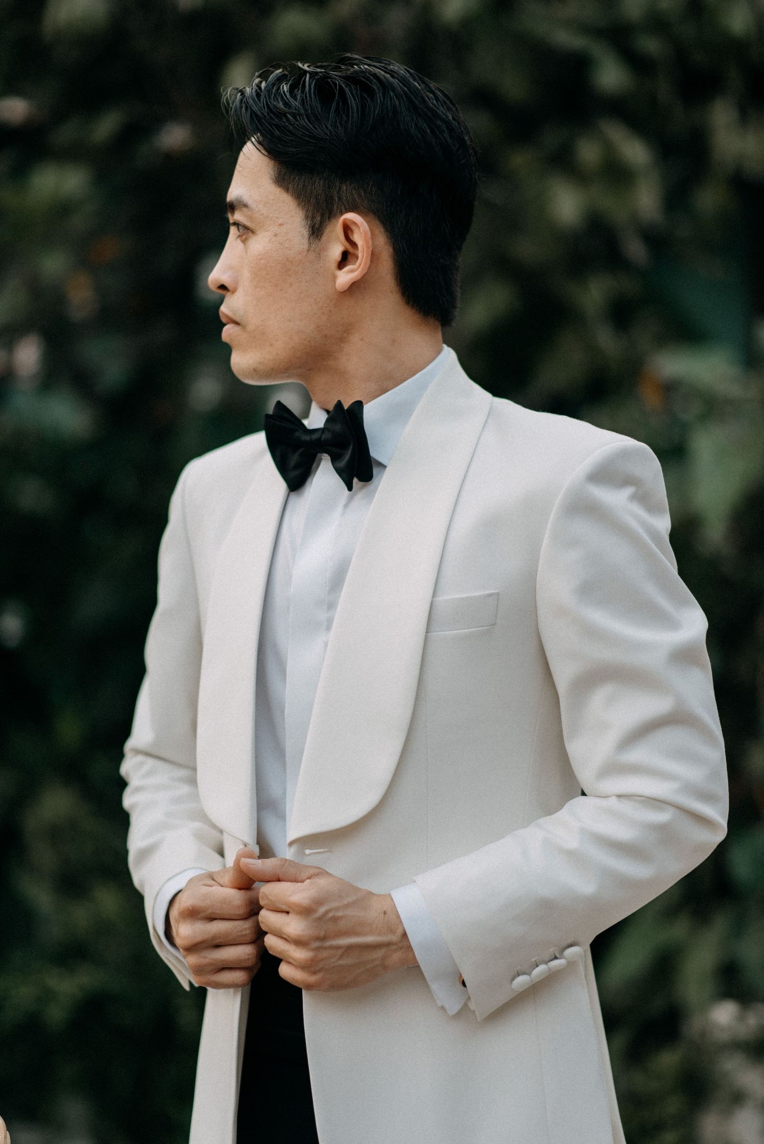 Side profile of person in white tuxedo jacket and black bow tie against green foliage.
