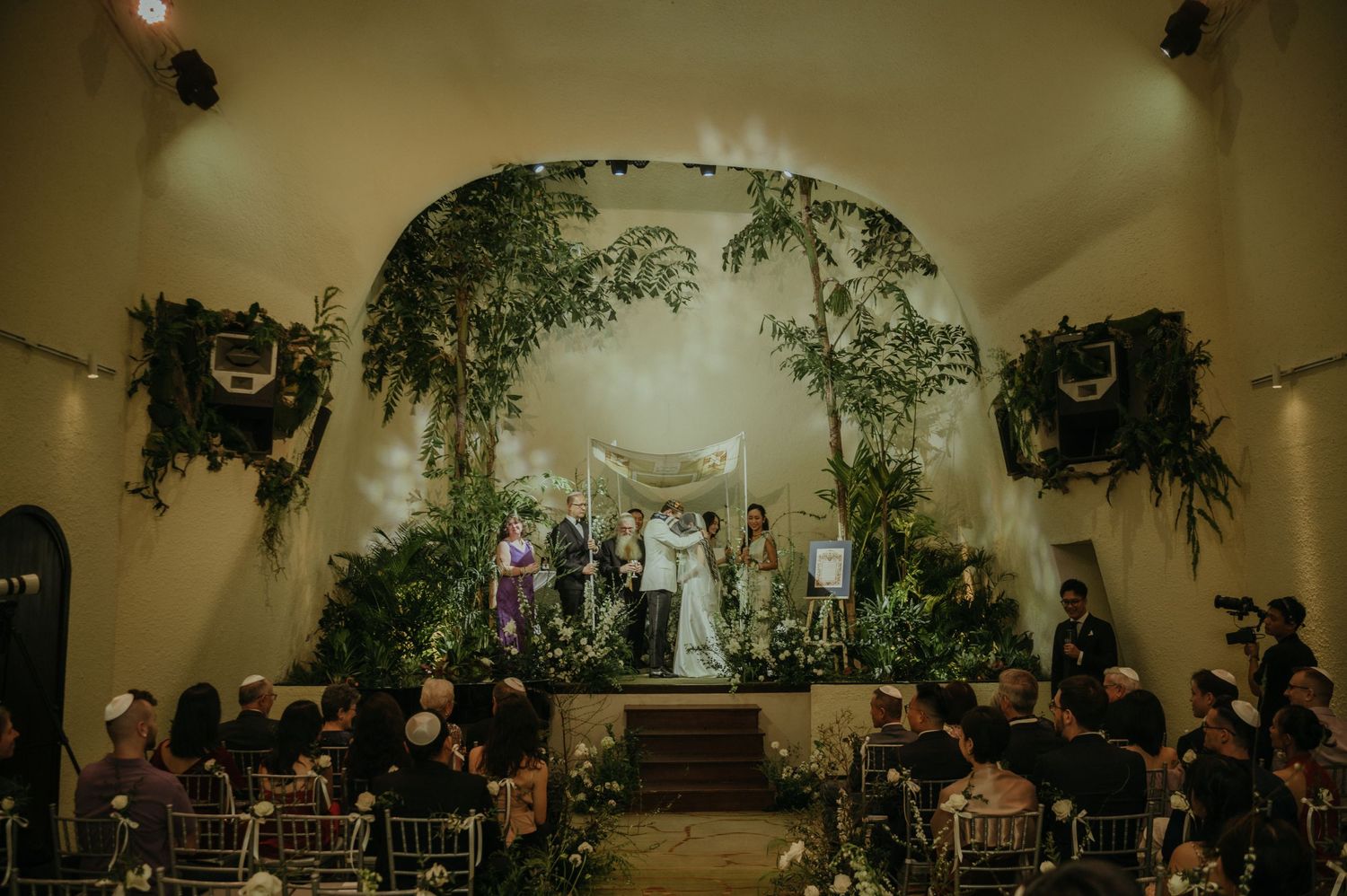 Wide-angle view of a wedding ceremony in a venue with arched ceiling and hanging greenery.