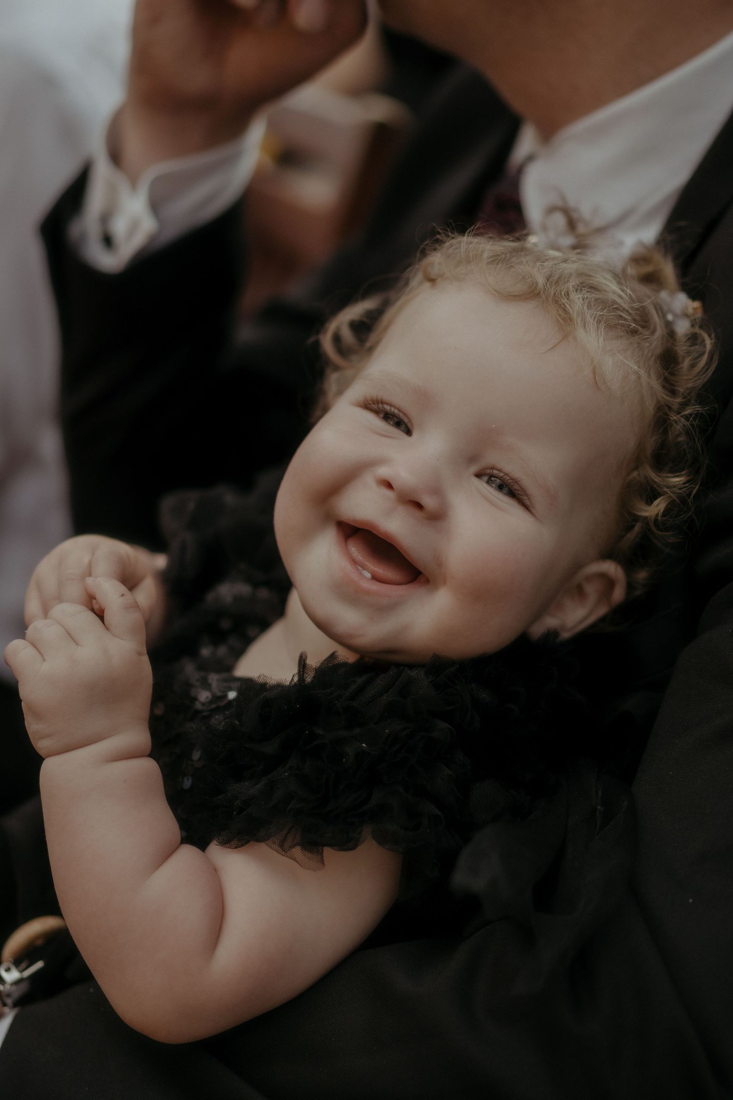A joyful baby in a black tulle dress smiles and gestures with raised hands during a formal occasion.
