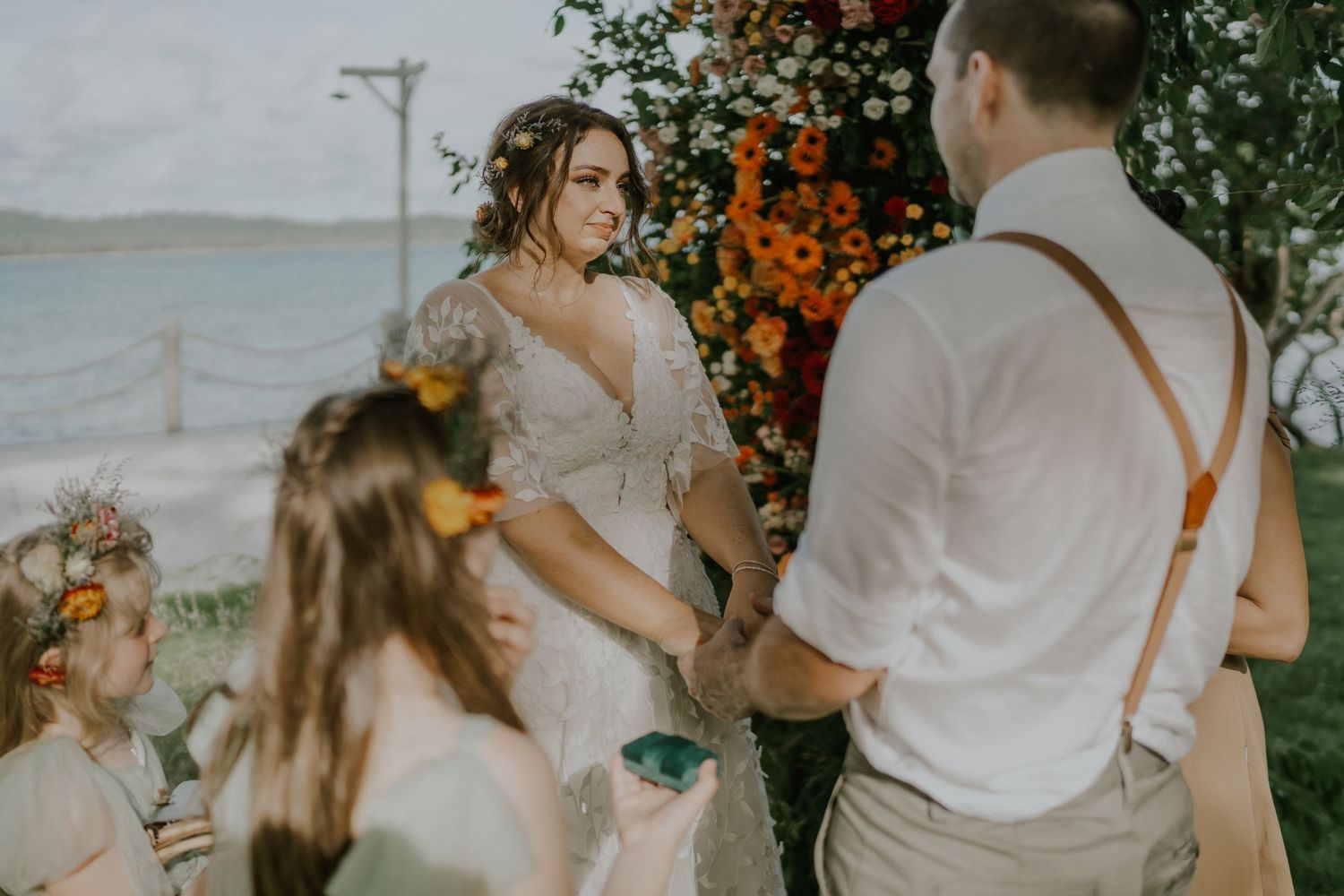 Outdoor waterfront wedding ceremony under floral arch with orange blooms.