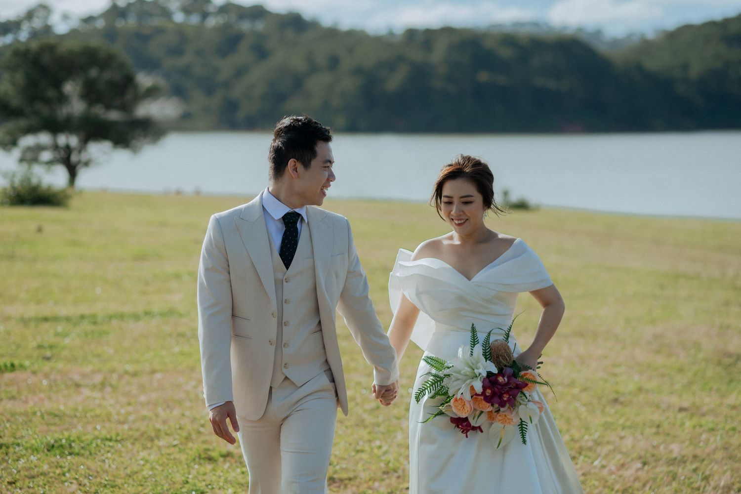 A couple in wedding attire walks on a grassy hill overlooking a serene lake, with the bride holding a colorful bouquet.