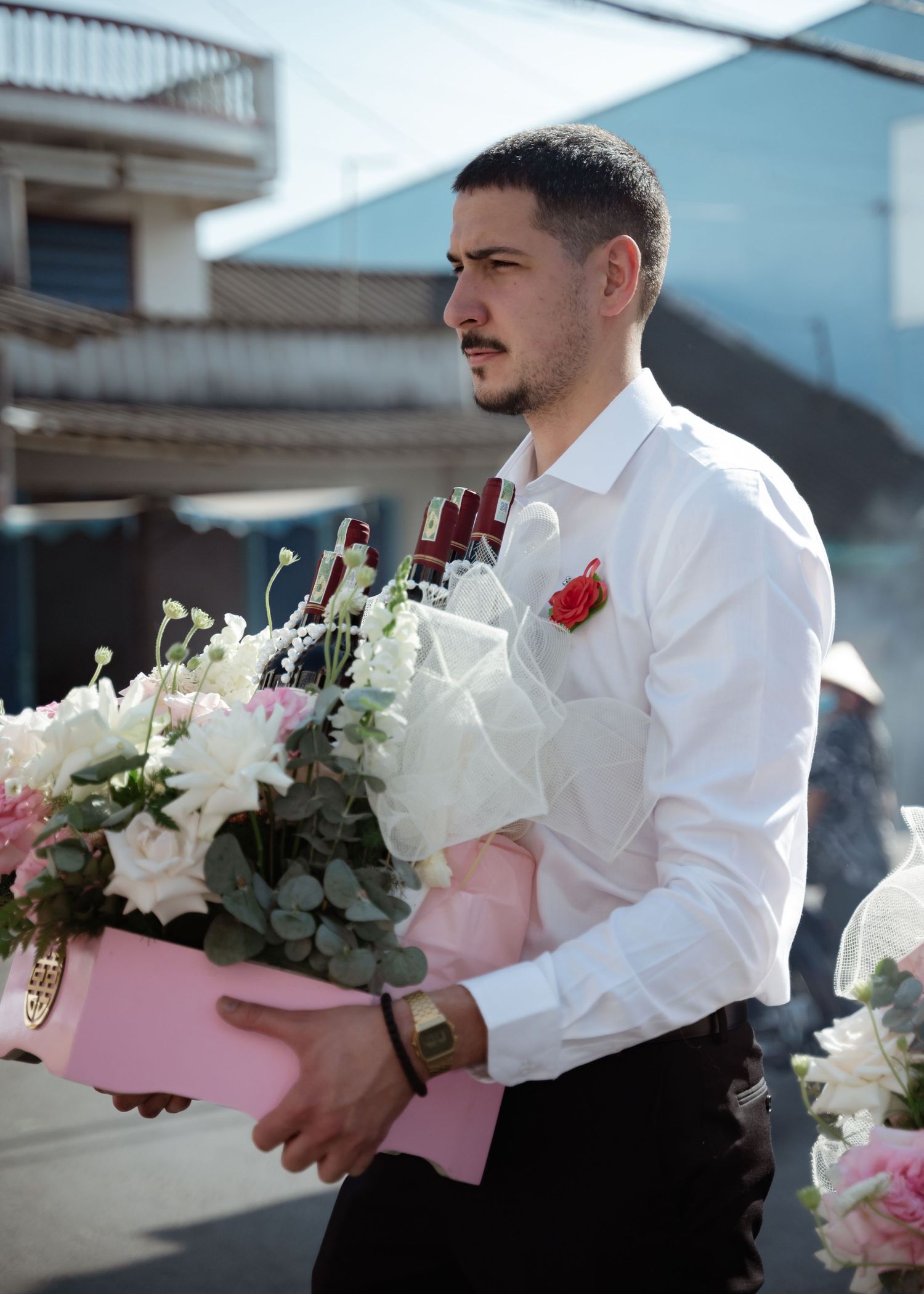 A man in a white dress shirt and red boutonniere carries a large pink and white floral arrangement outdoors.