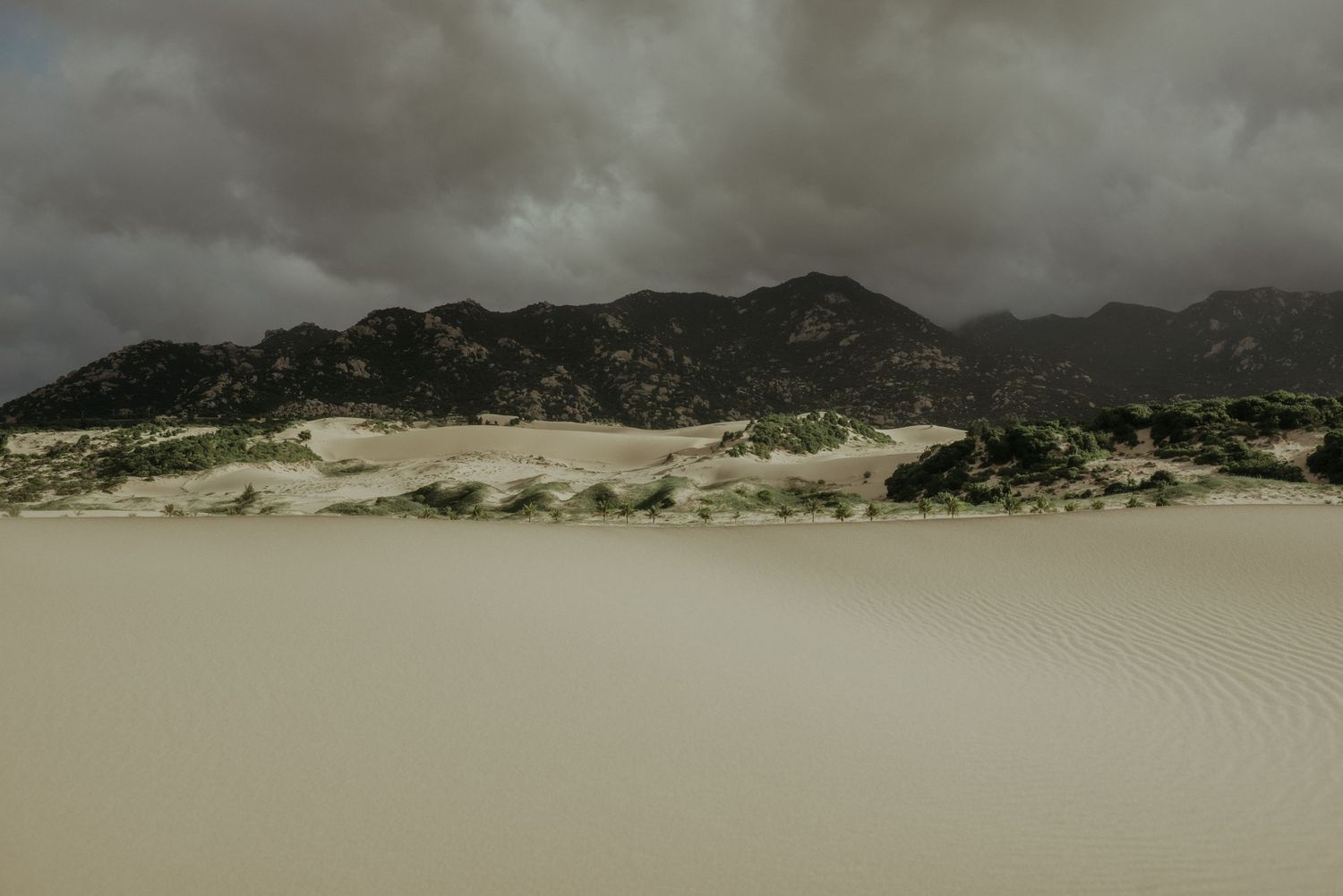 Dark storm clouds loom over sand dunes and rocky mountains in a dramatic desert landscape.