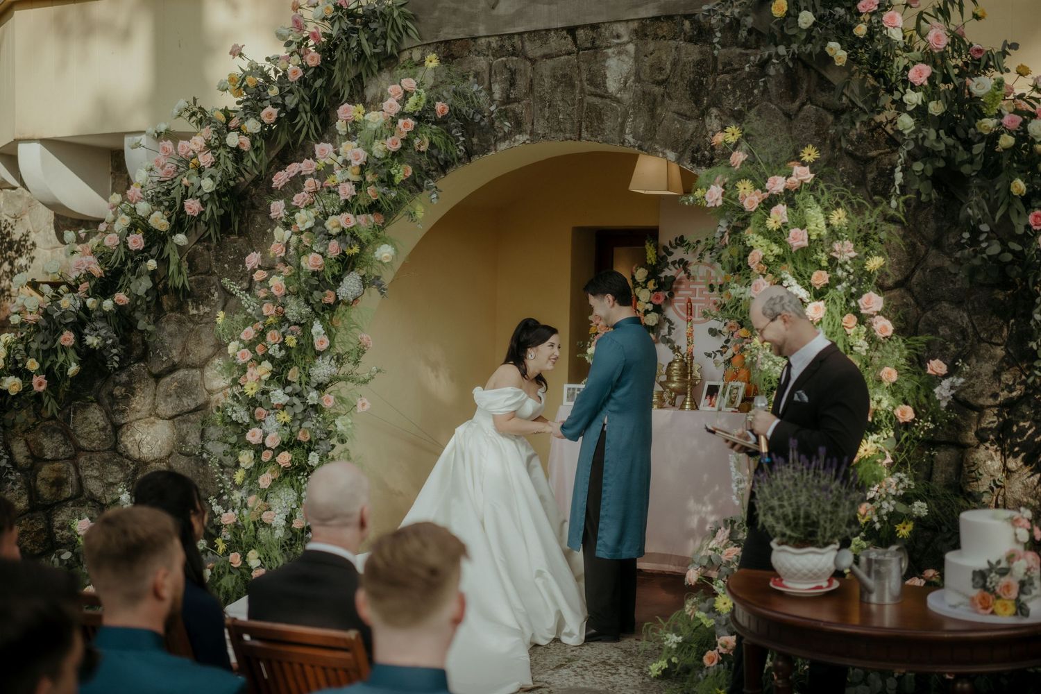 Wedding ceremony in a romantic stone archway decorated with climbing roses and floral arrangements in a garden setting.