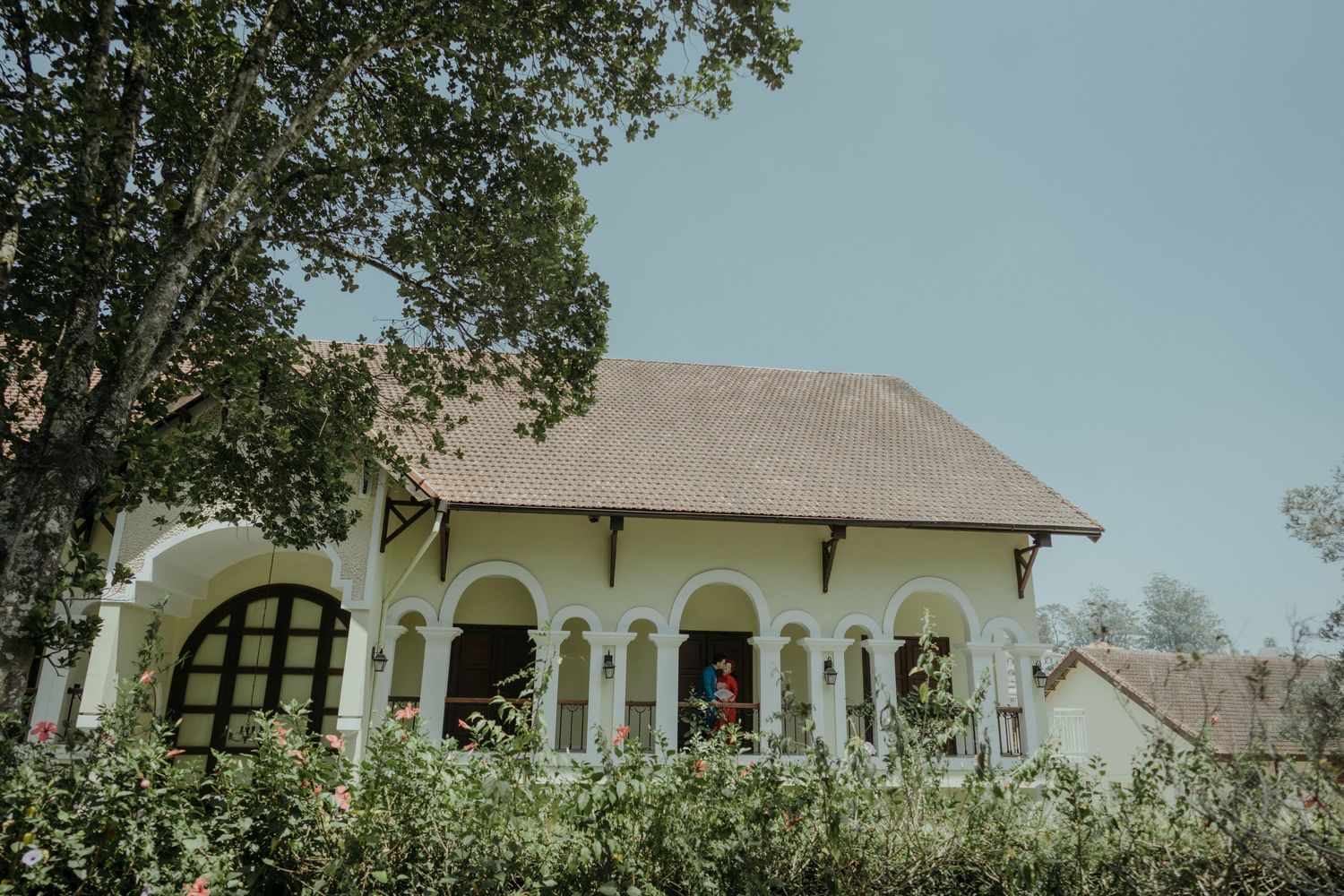 Vintage white villa with arched windows and covered porch surrounded by lush greenery against blue sky.