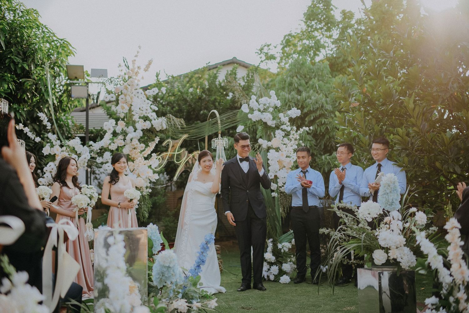 Outdoor wedding ceremony under floral arch with guests seated on white chairs.