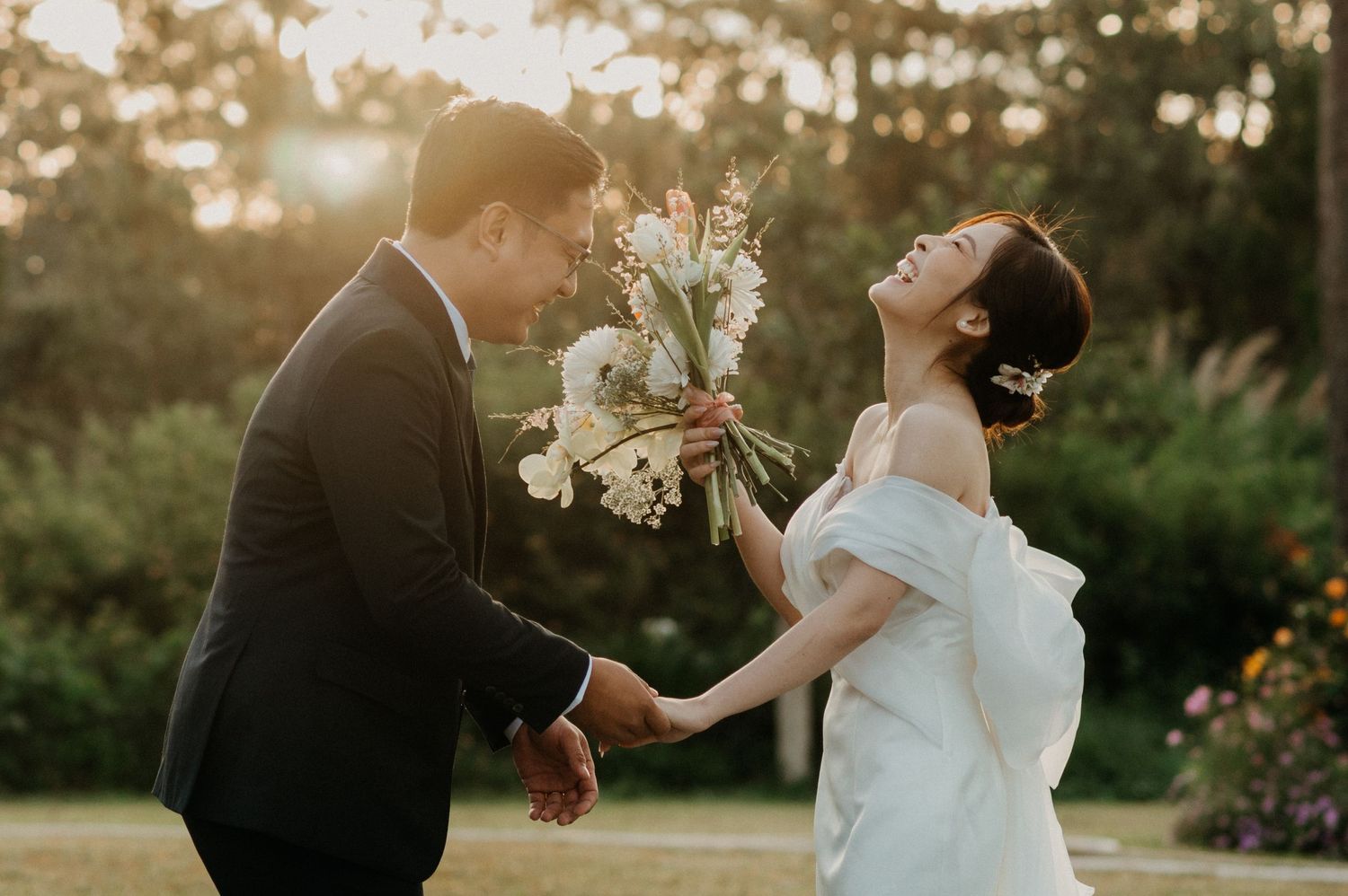 A joyful wedding couple laughs together while holding a white bouquet during golden hour in a garden setting.