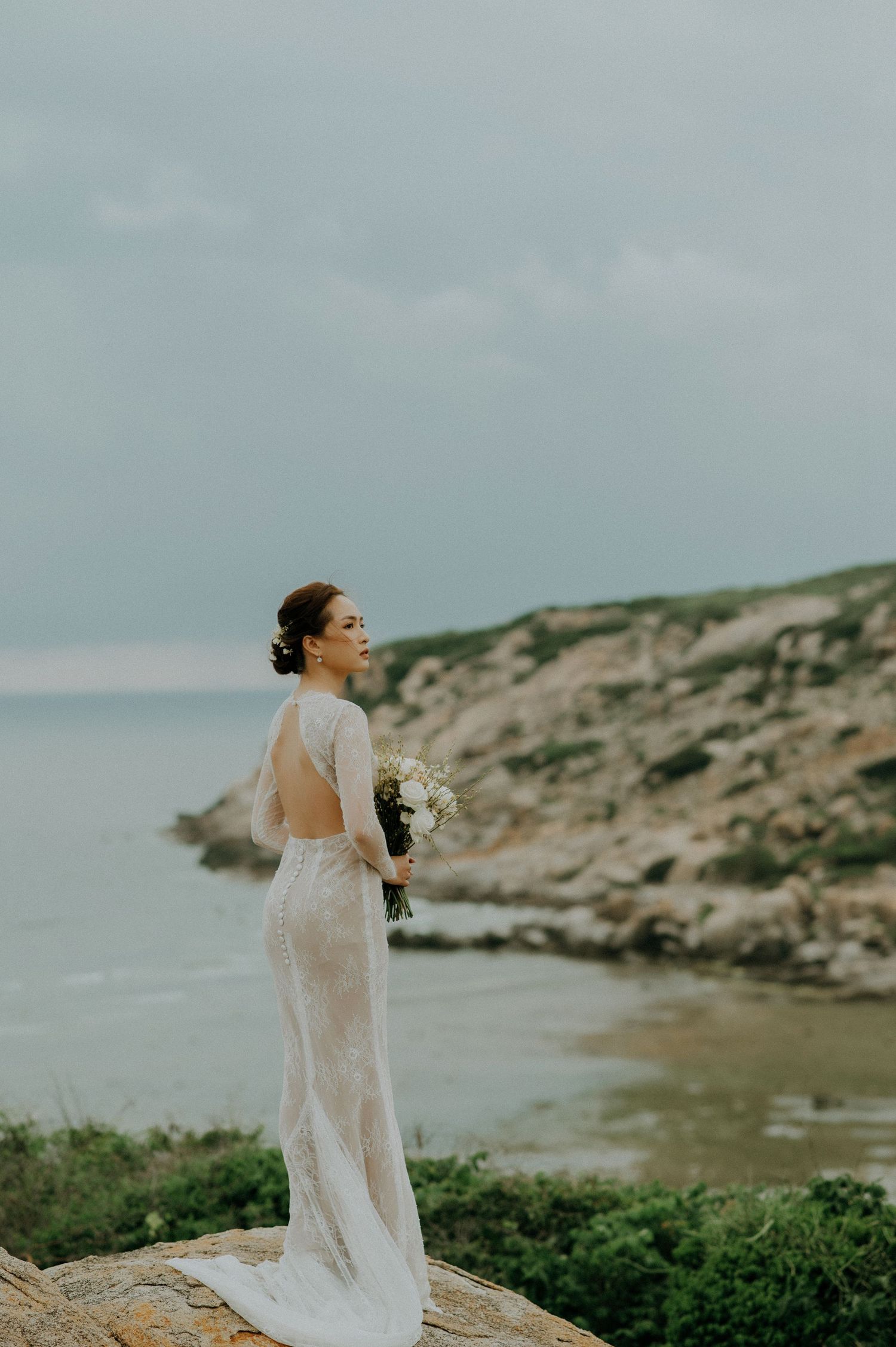 A bride in a sheer lace gown holds a white bouquet while standing on coastal cliffs overlooking the ocean.