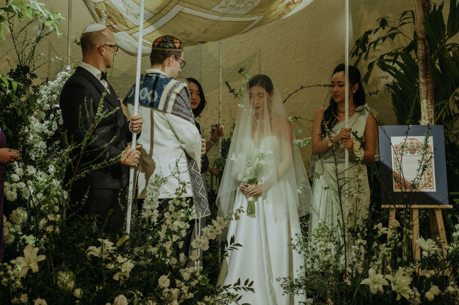 A wedding ceremony taking place on a stage decorated with white flowers and greenery under a draped canopy.