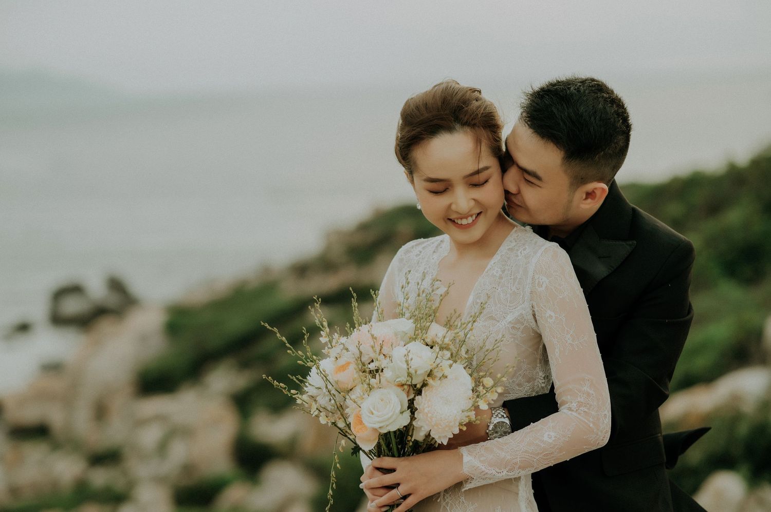 A loving couple shares an intimate moment on a coastal cliff while holding a delicate bouquet of white flowers.