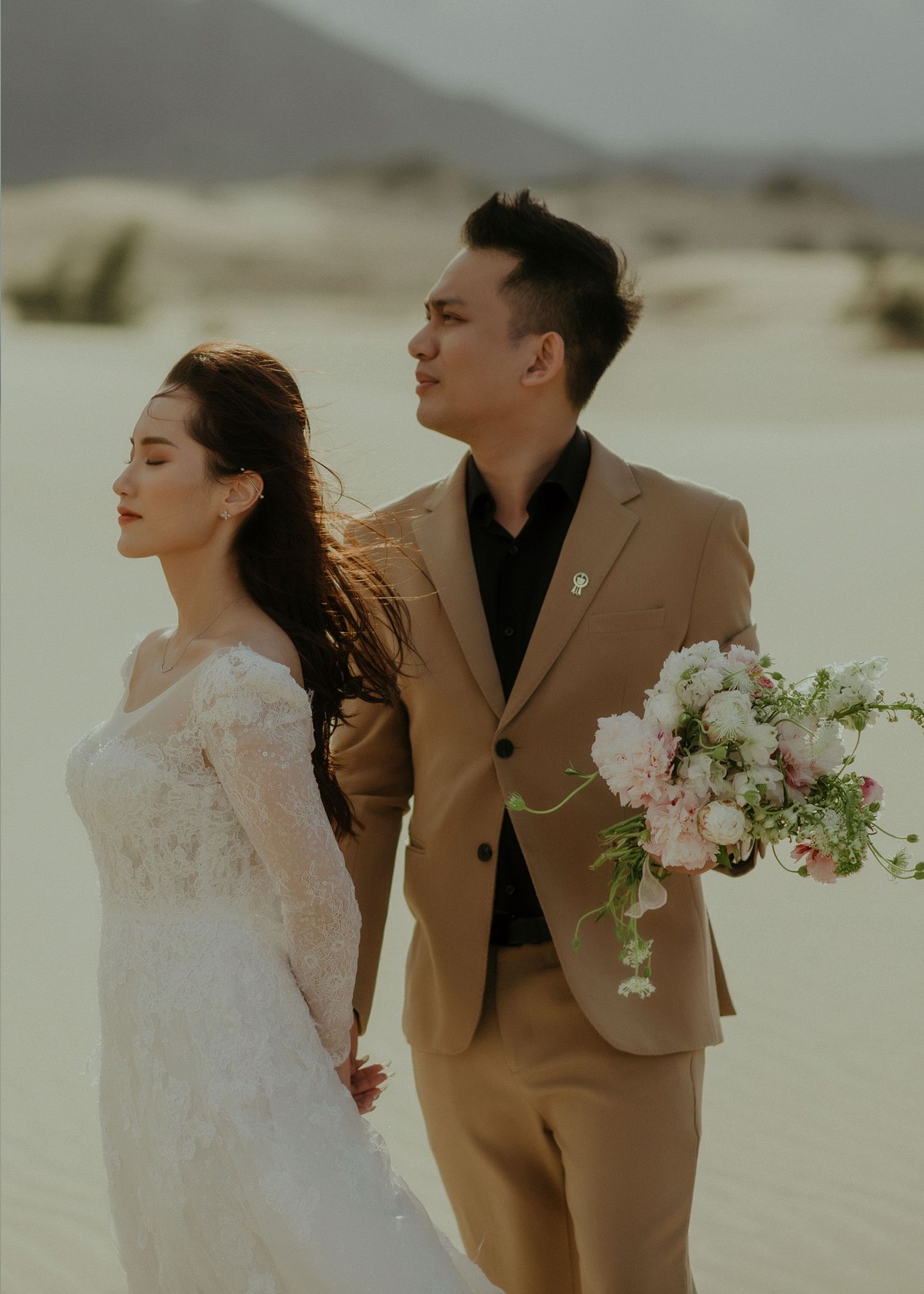 A couple in wedding attire stands against desert sand dunes, with the bride holding a soft pink floral bouquet.