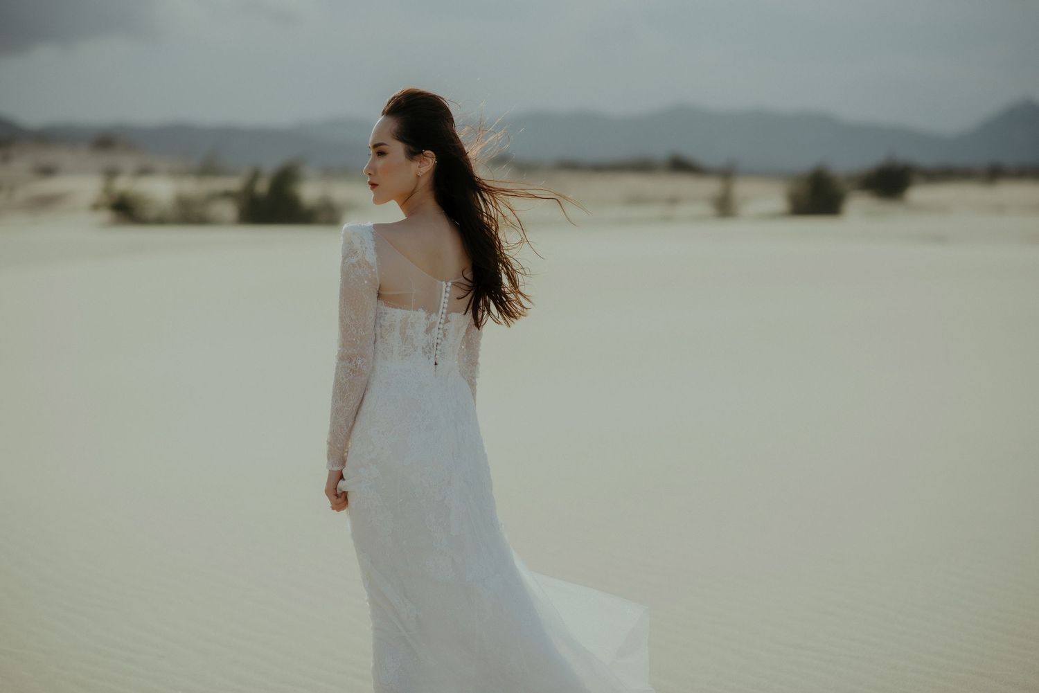 A bride in a lace white dress stands alone in a desert landscape with wind blowing through her hair.