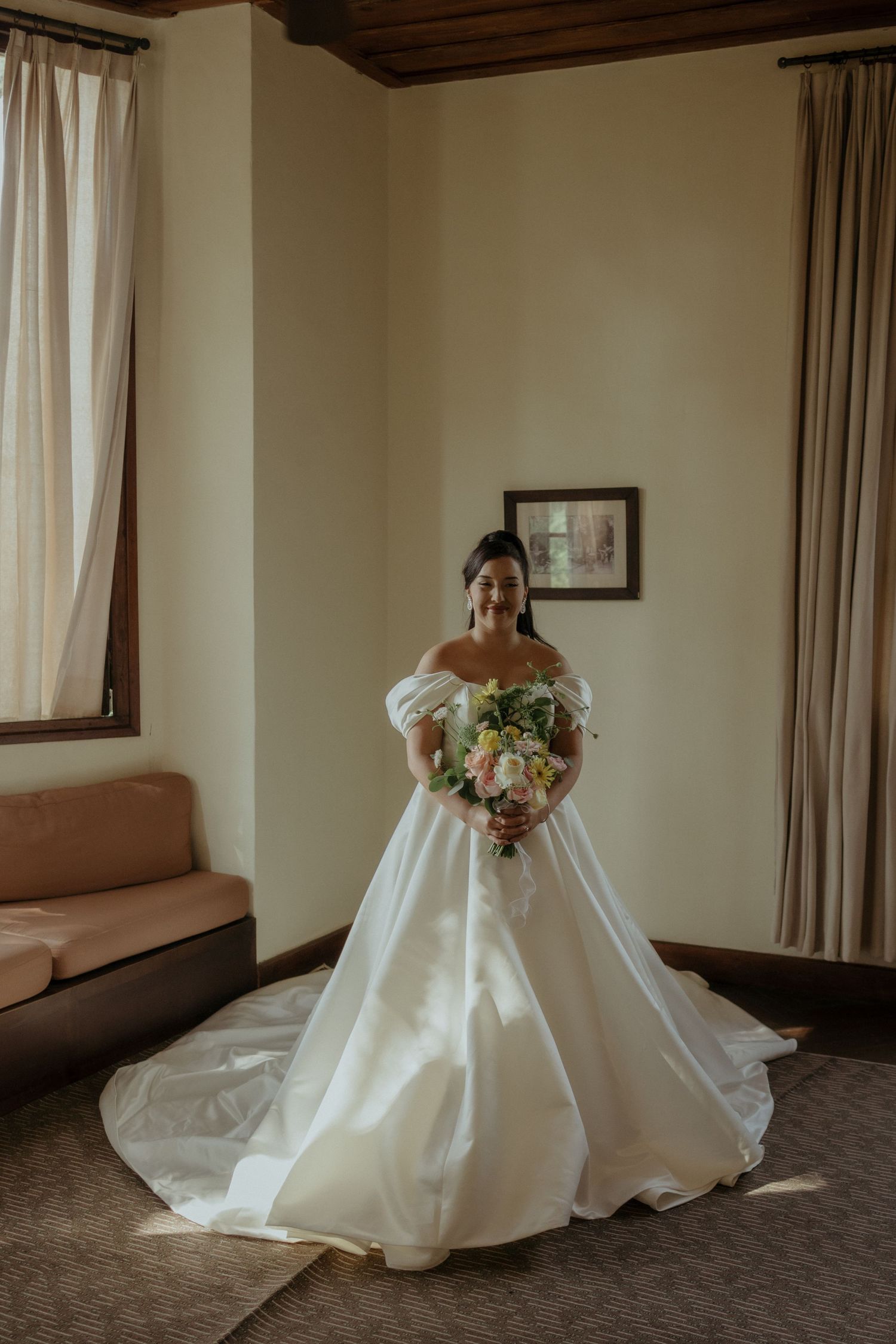 A bride in an elegant white off-shoulder wedding gown holding a pastel bouquet stands in a room with curtained windows.