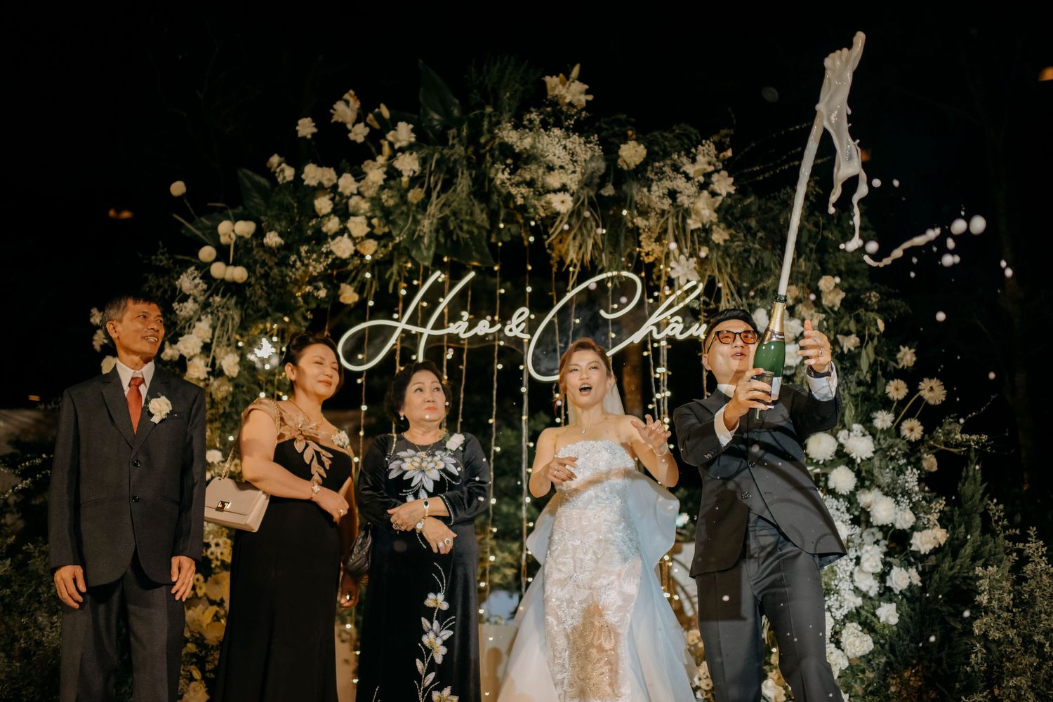 Wedding party poses under elegant floral arch with illuminated script signage at night.