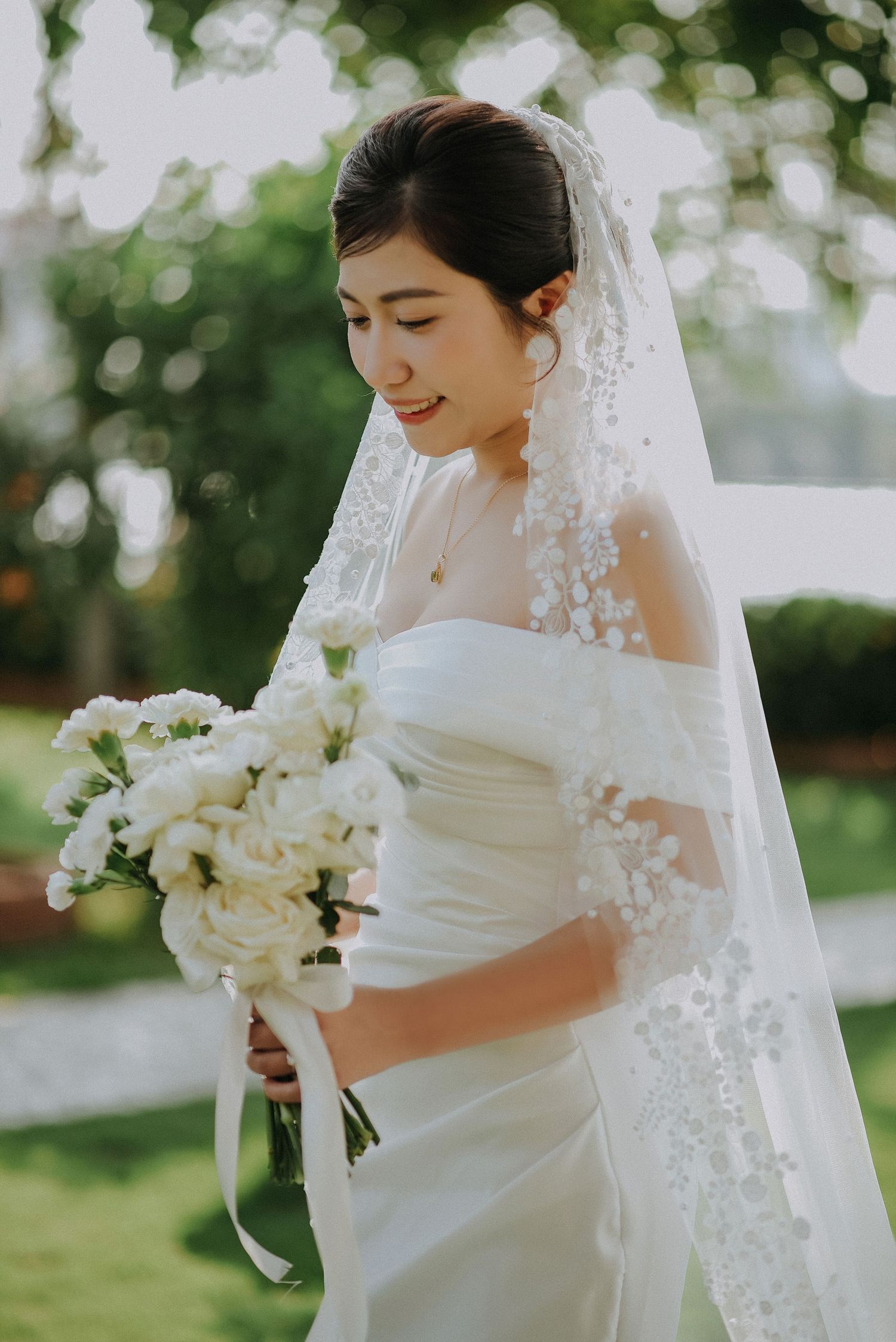 Elegant bride in white off-shoulder dress holding classic white bouquet during outdoor ceremony.