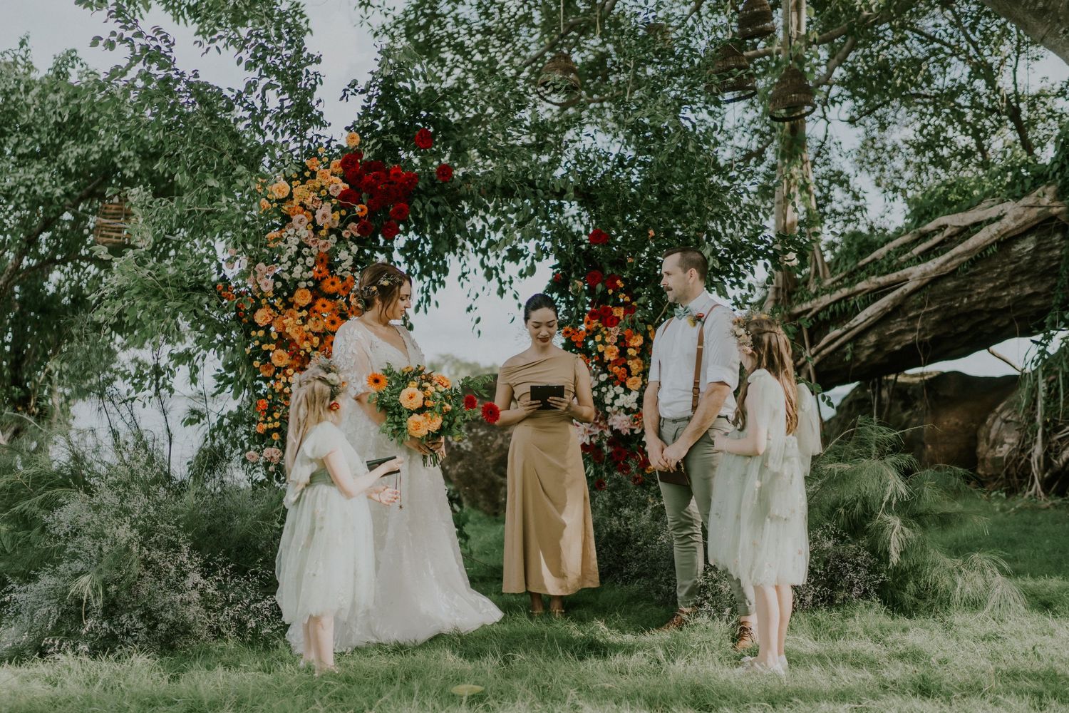 Wedding party standing under floral arch with orange and yellow flowers in outdoor garden setting.