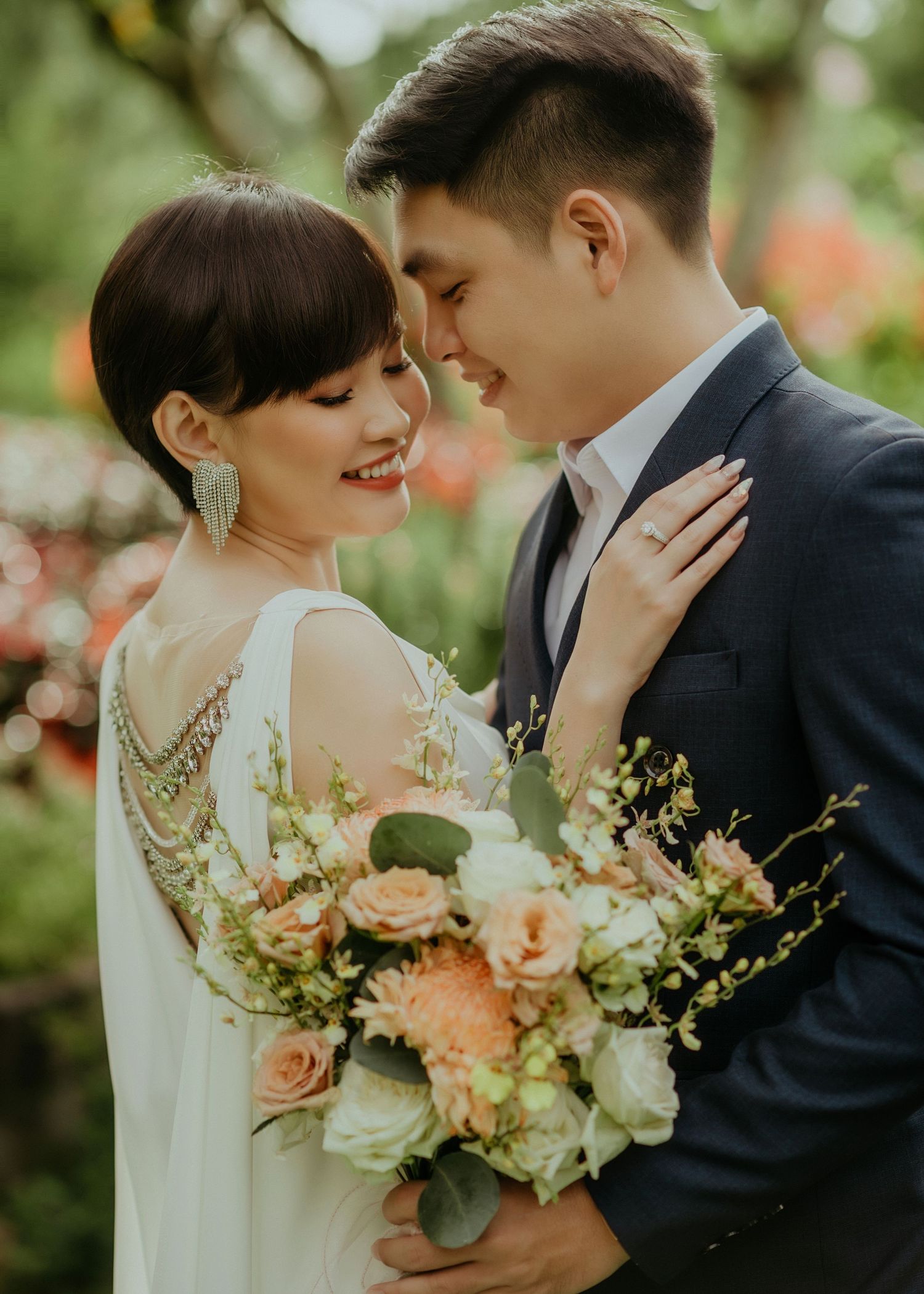 Romantic wedding couple shares an intimate moment in a garden with a beautiful peach and white floral bouquet.