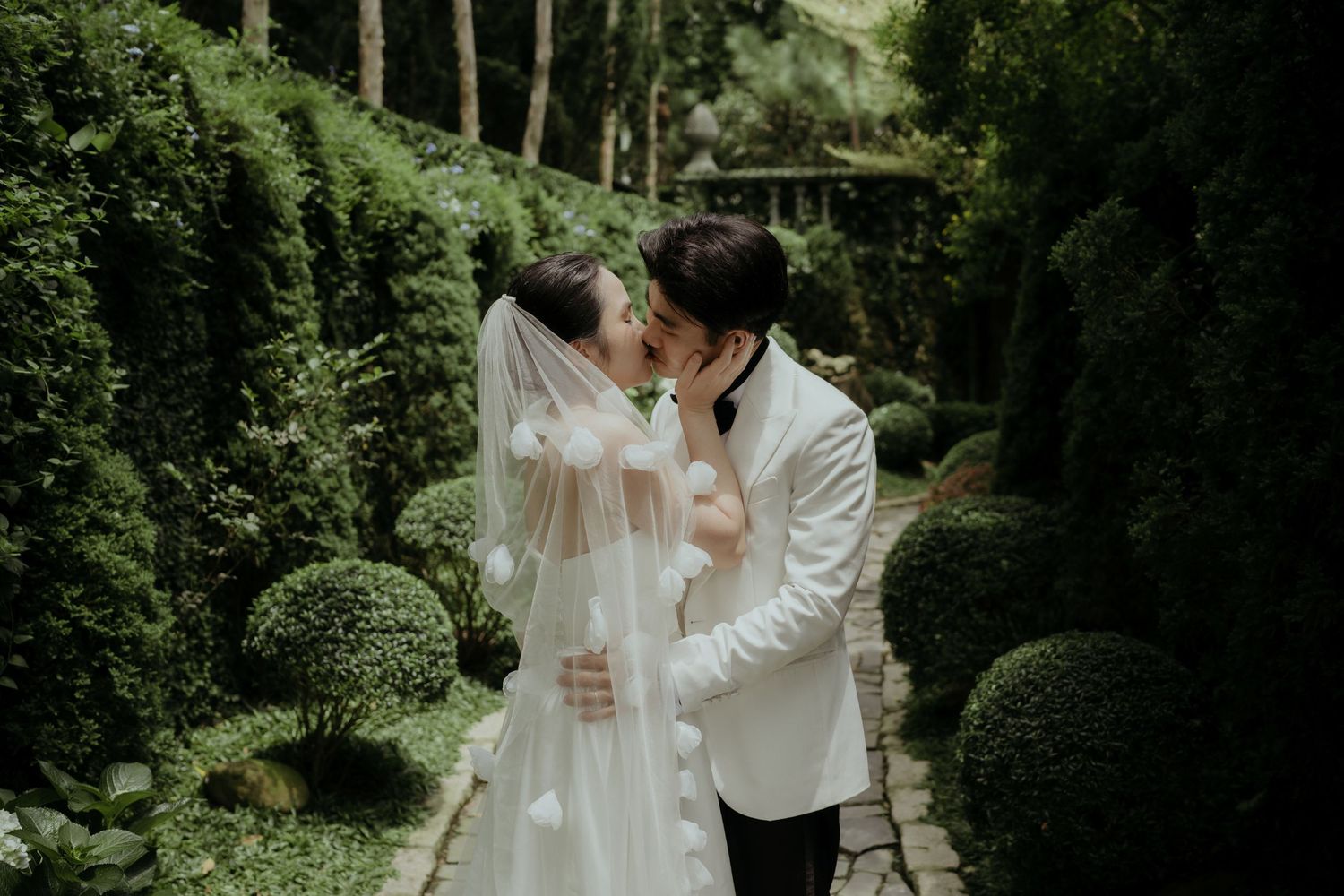 A couple embraces on a romantic garden path surrounded by lush greenery and manicured shrubs on their wedding day.