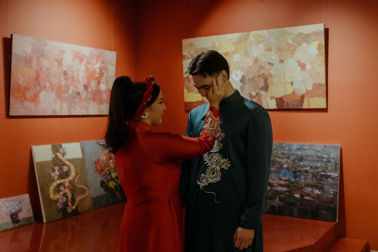 Two people share an intimate moment in front of abstract artwork displayed on orange-colored walls in a gallery setting.