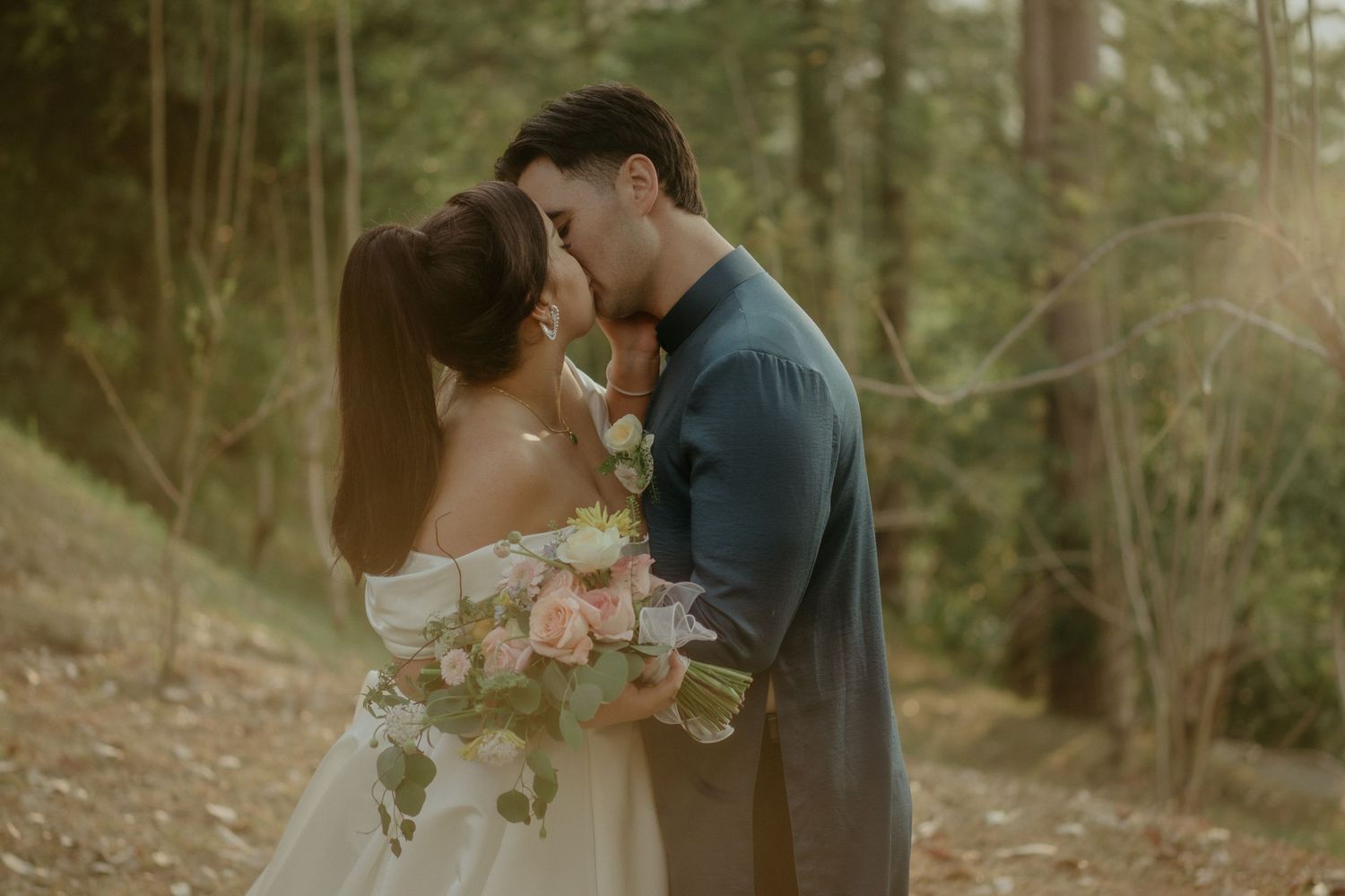 A romantic couple shares an intimate moment in a forest while holding a bouquet of blush roses.