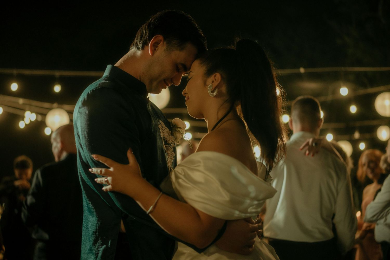 A couple shares a romantic dance moment under string lights at an outdoor evening celebration.