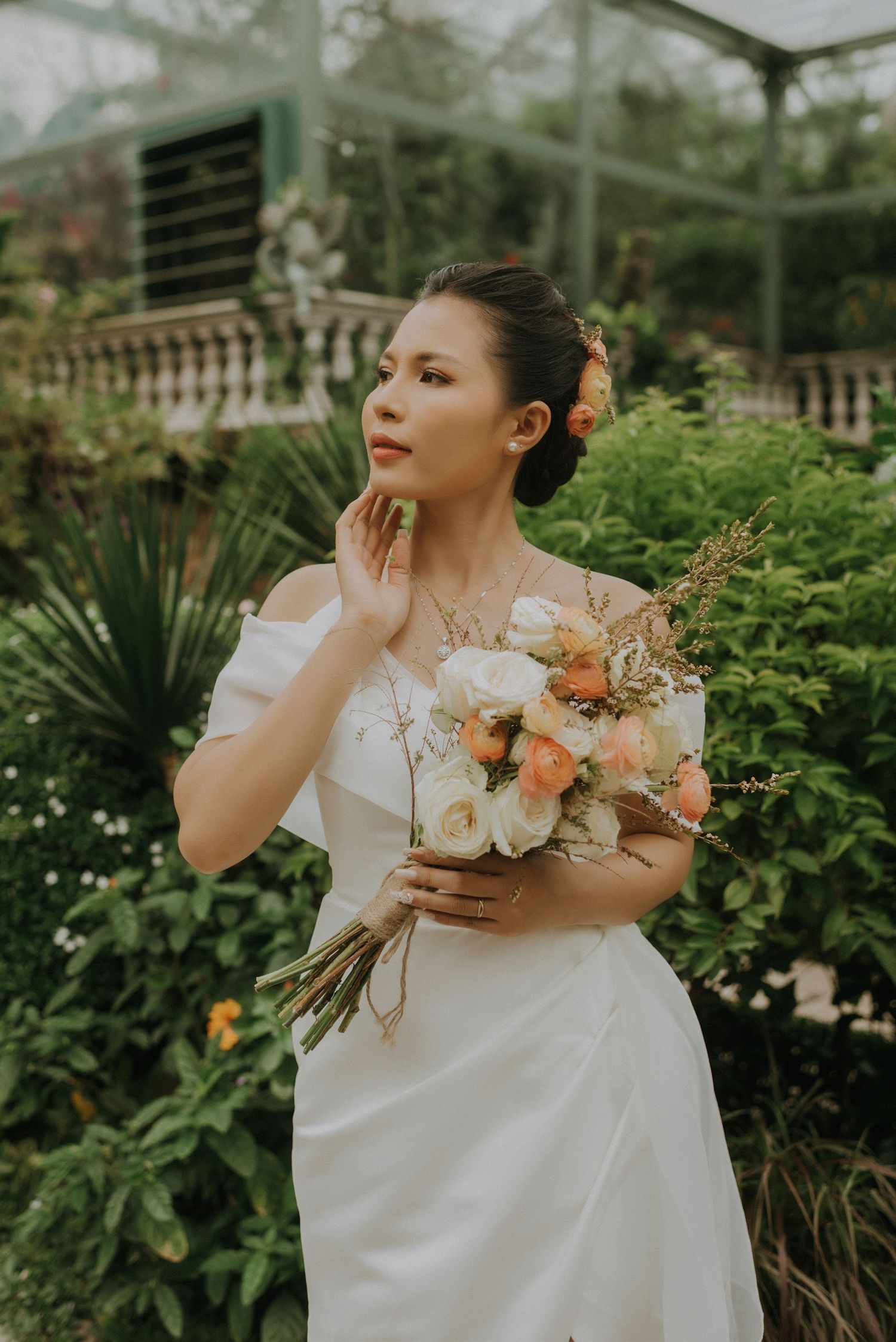 Elegant bride in off-shoulder white dress poses with peach and white floral bouquet in a lush garden greenhouse setting.