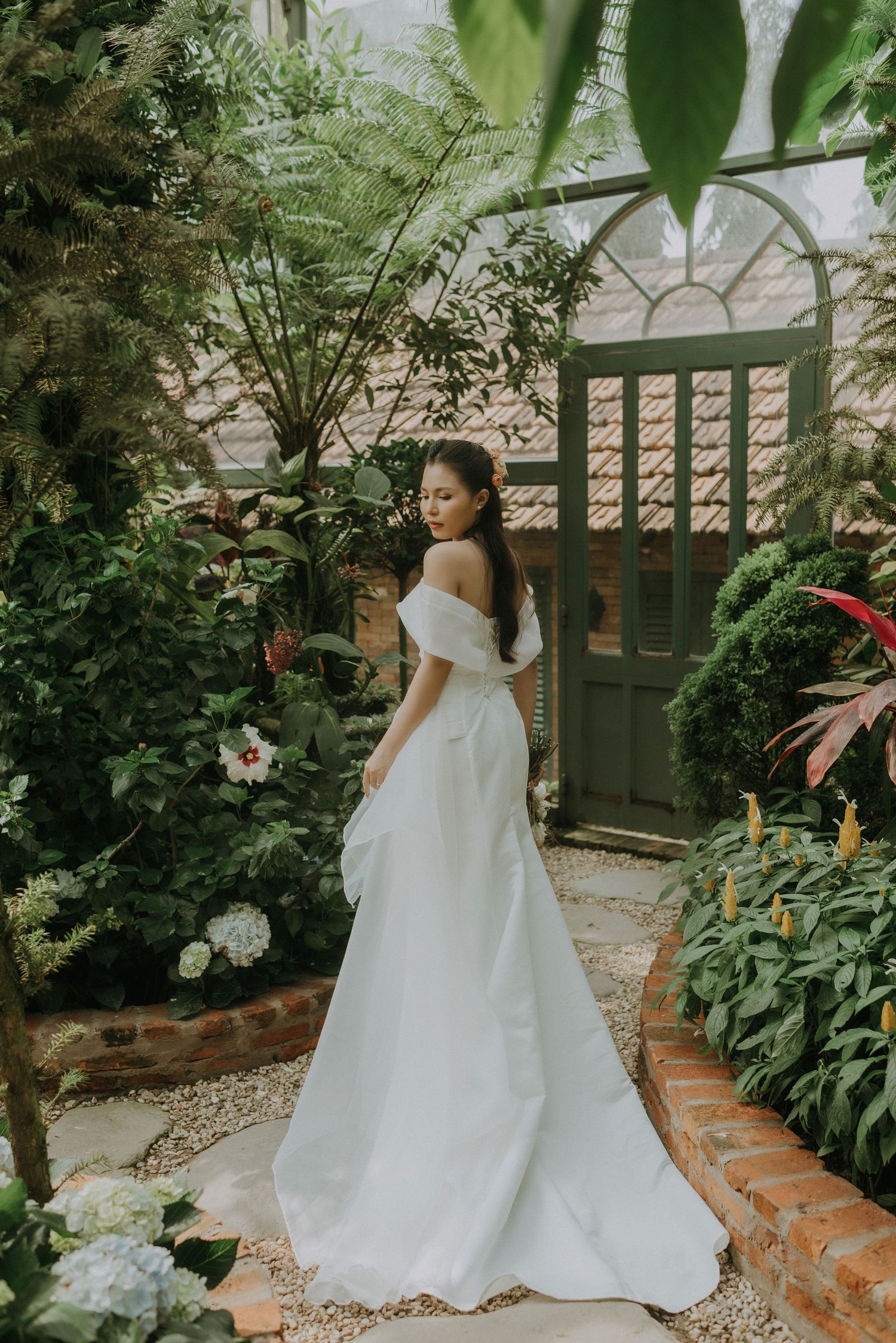 A person in an elegant white off-shoulder wedding dress poses in a lush garden setting with brick steps and wooden gate.