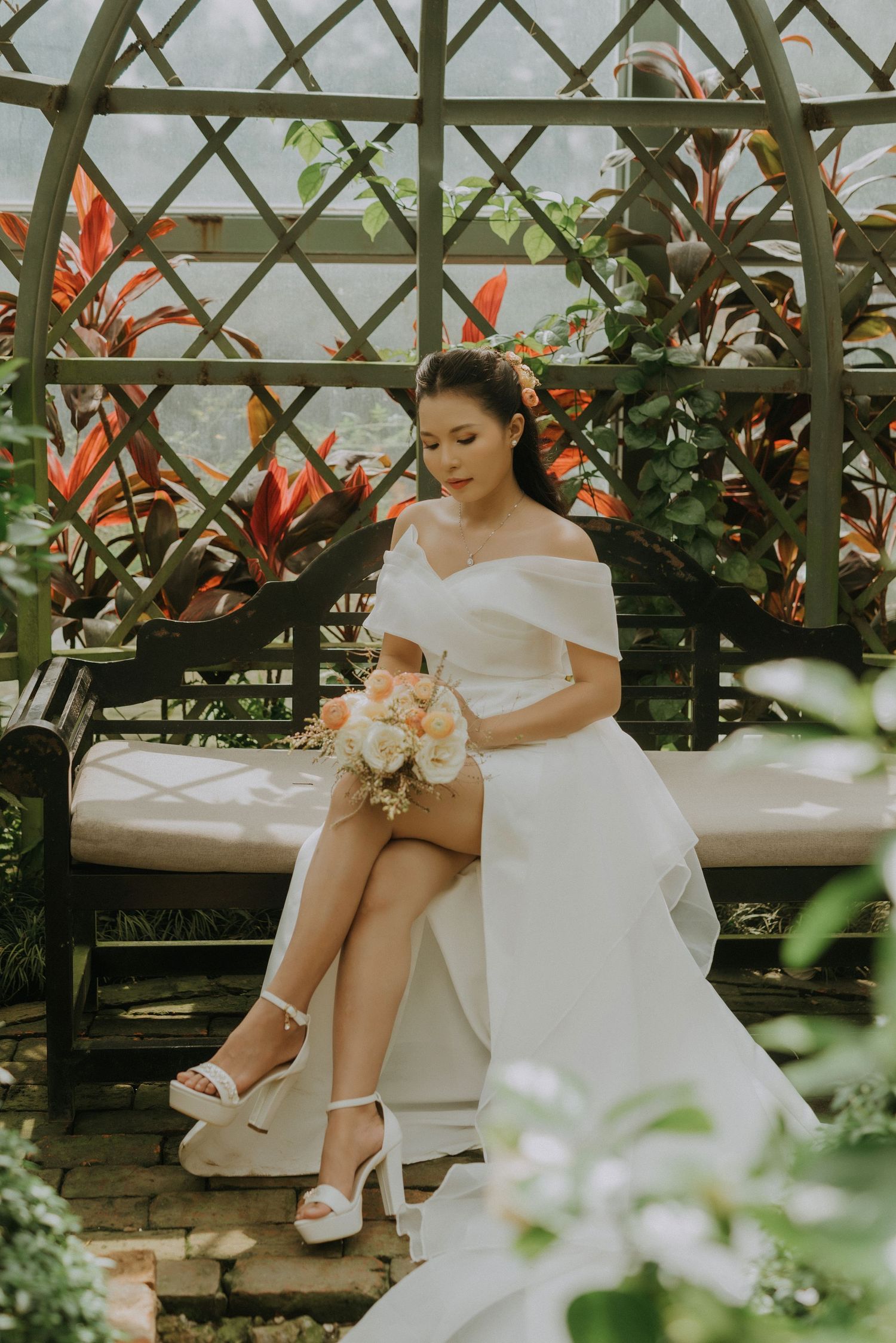 A bride in an off-shoulder white wedding dress sits elegantly in a greenhouse surrounded by tropical foliage and flowers.