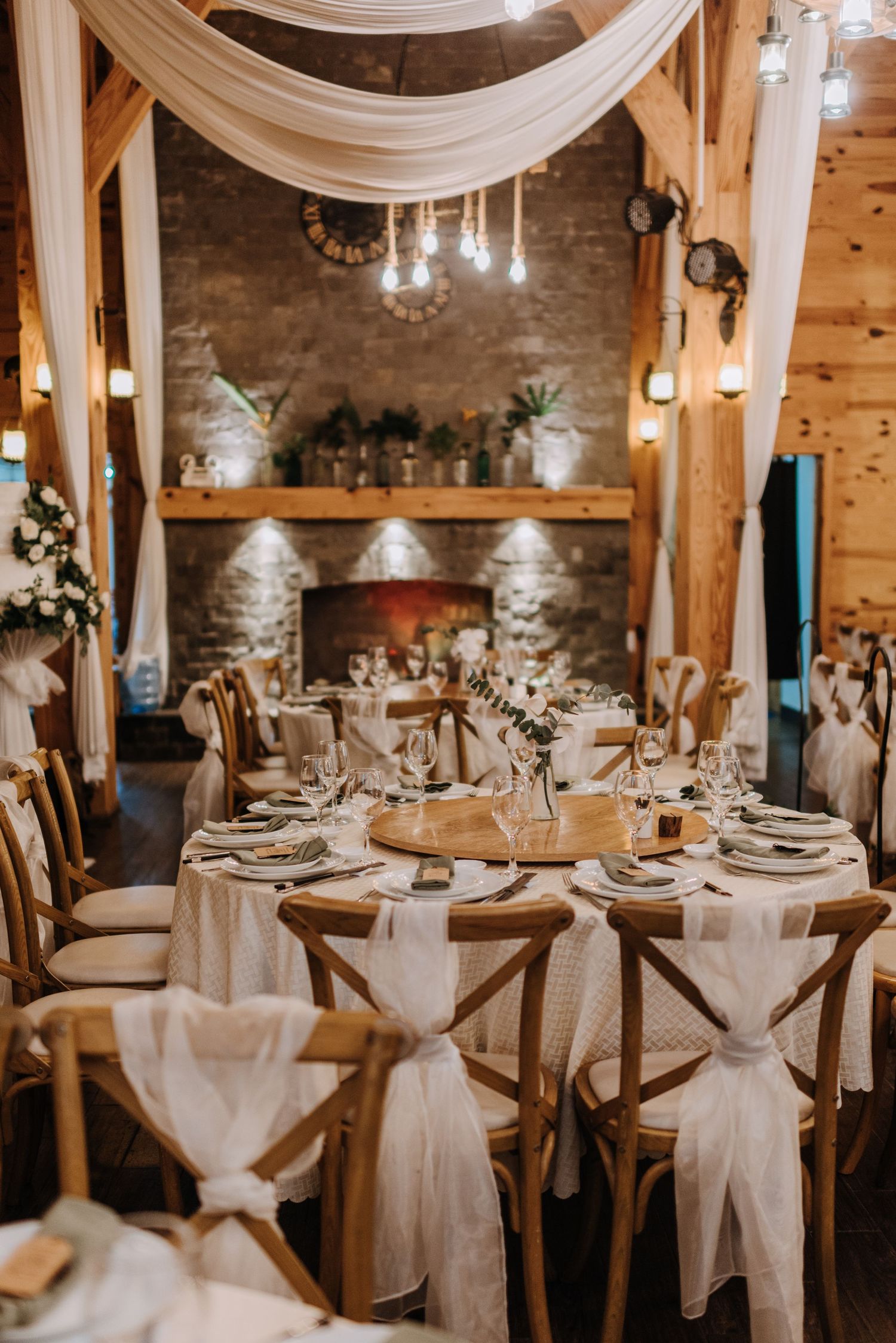 An elegant wedding reception setup with white draping, cross-back chairs, and crystal chandeliers.