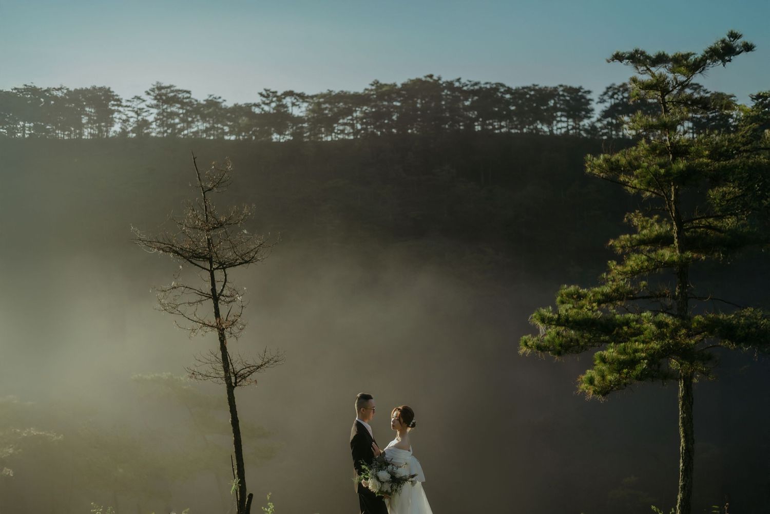 Two silhouetted figures stand between trees on a misty mountainside during a moody sunset in nature.