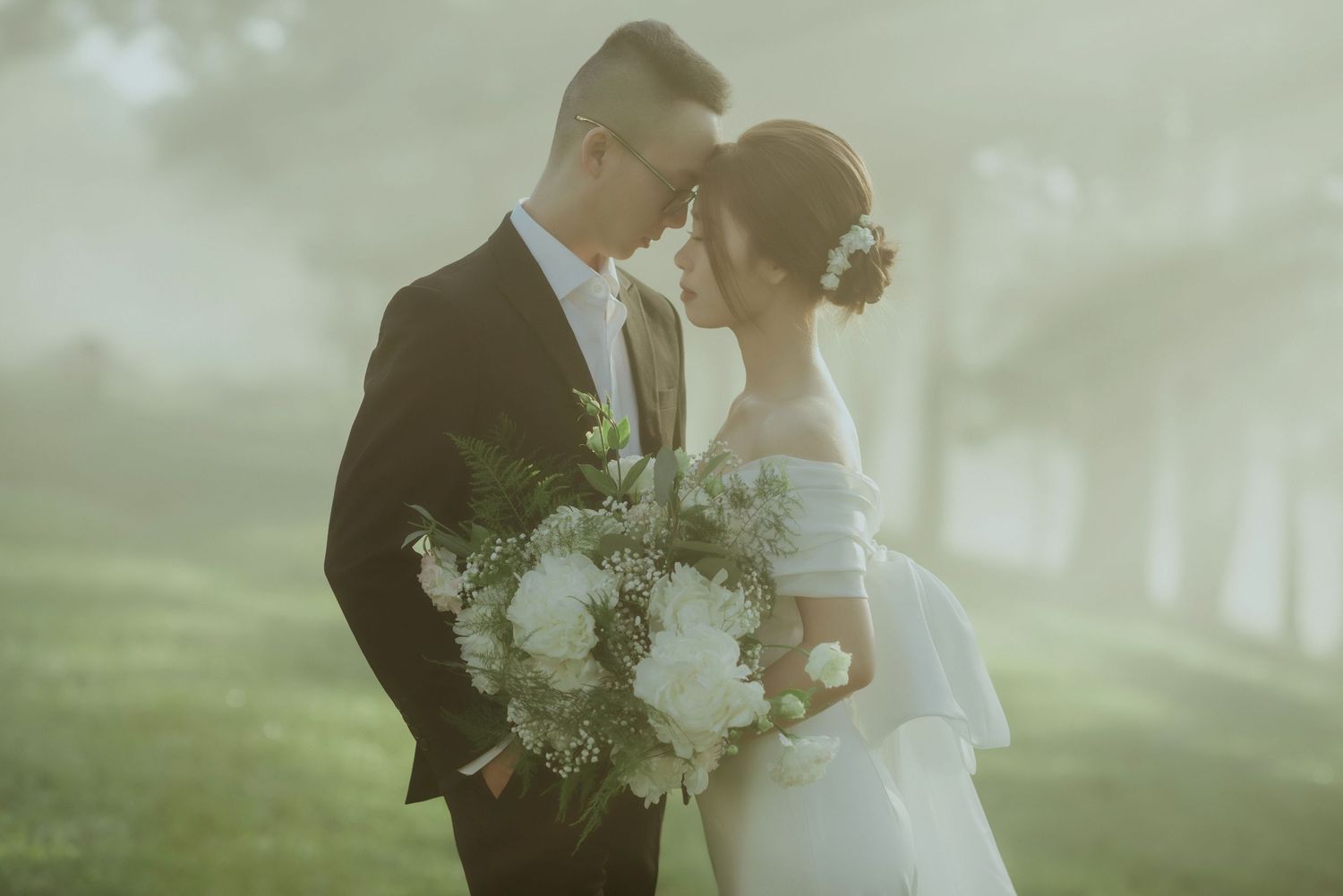 A romantic couple in wedding attire embrace in a misty garden setting while holding a white floral bouquet.